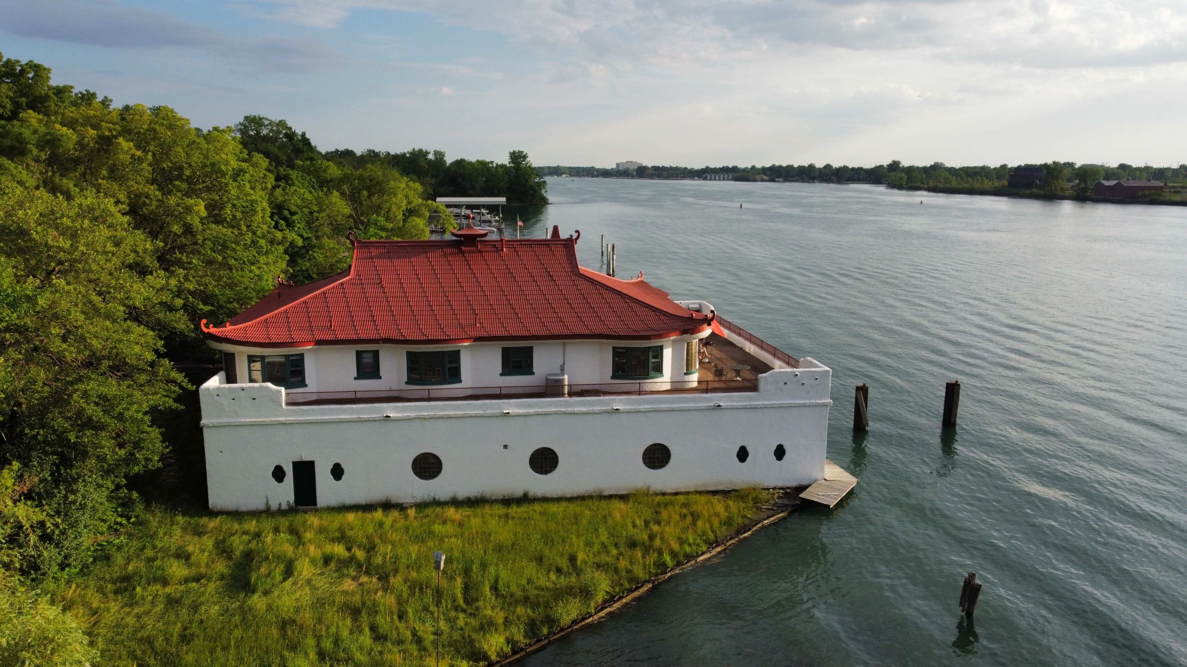 A white building with a red-tiled roof is situated on the edge of a river, surrounded by greenery and water.