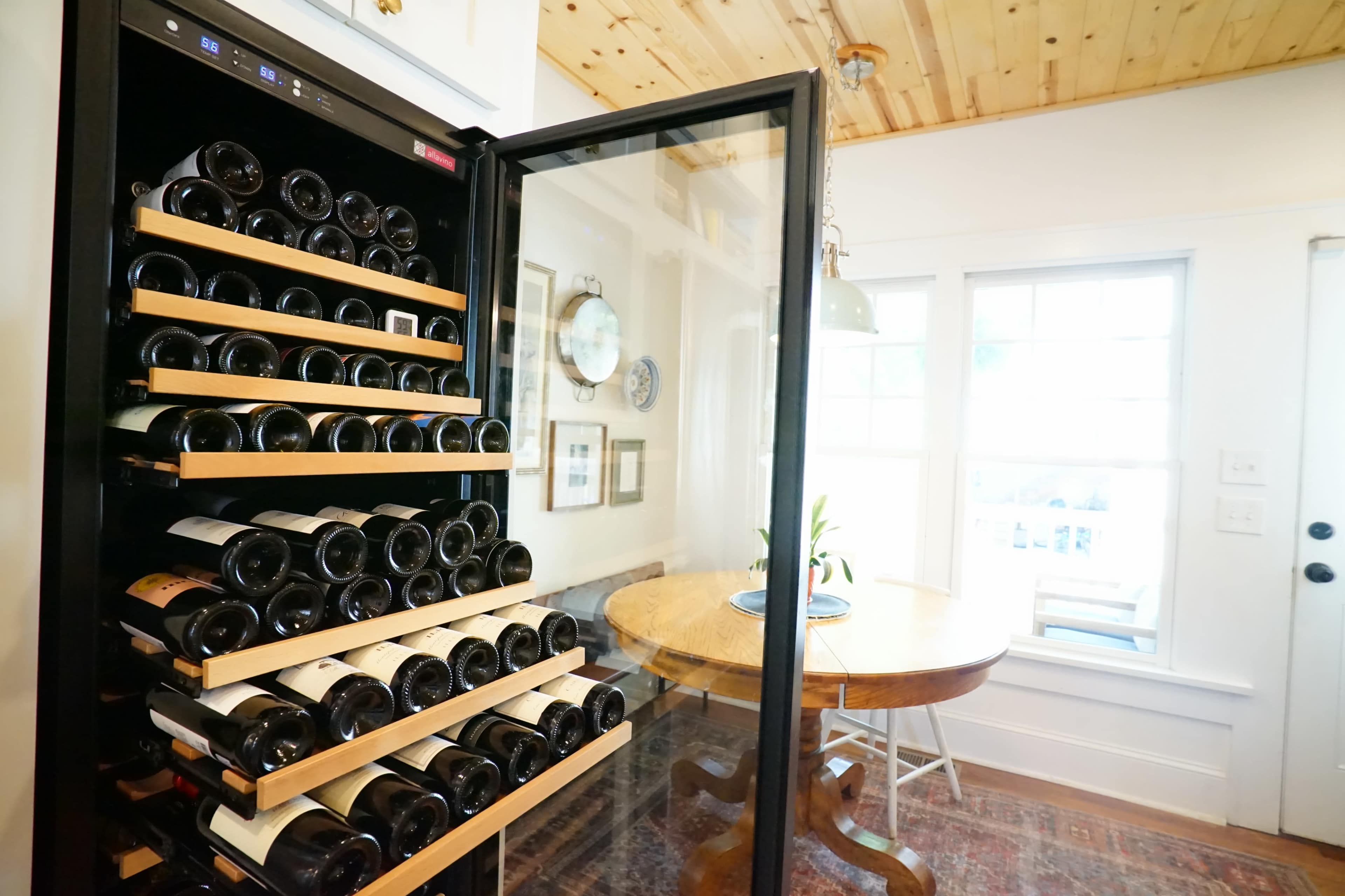 A wine refrigerator filled with bottles is positioned beside a round wooden dining table near a window in a well-lit room.