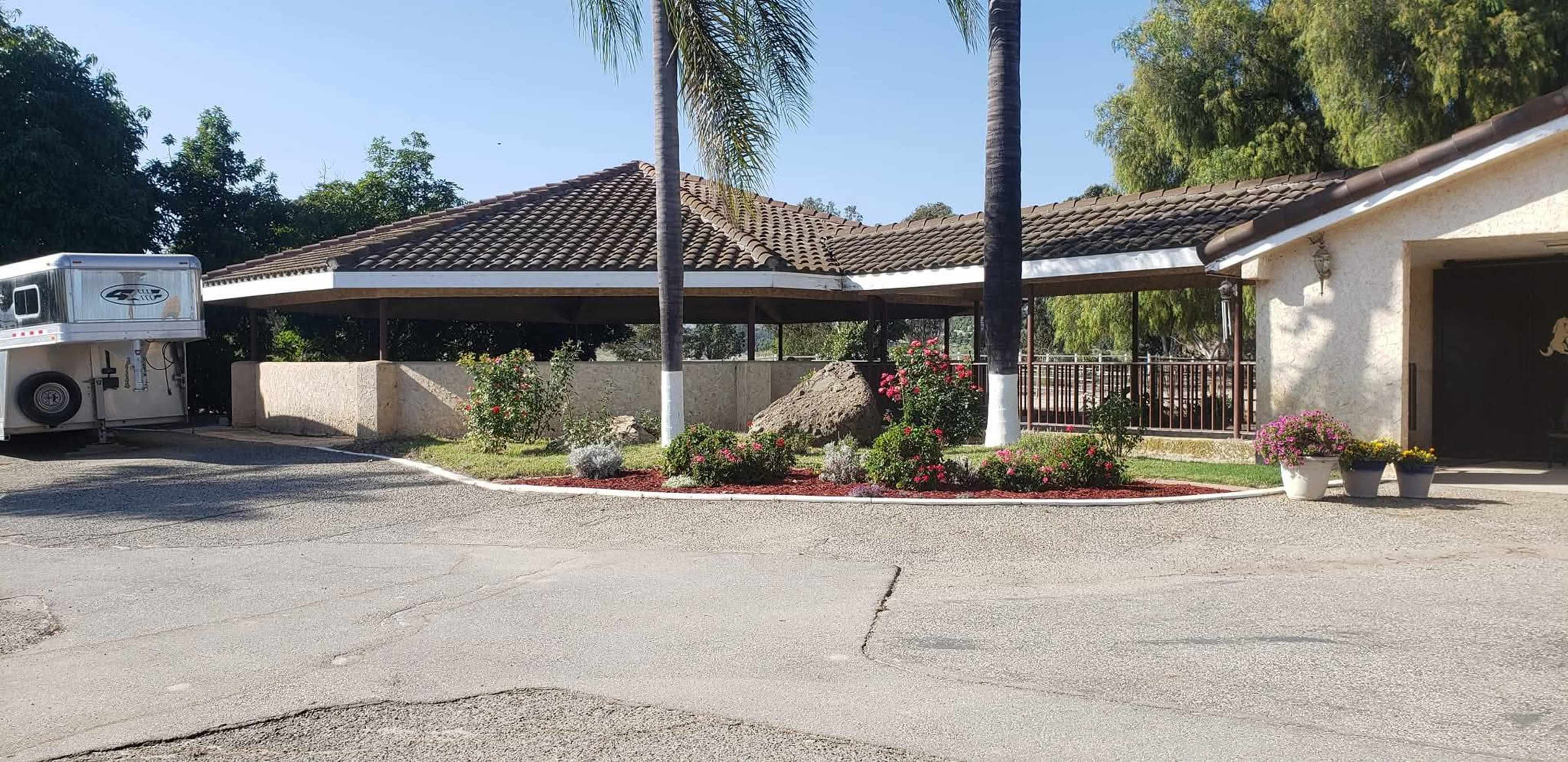 The image shows a residential property featuring a covered patio bordered by flower beds, with a horse trailer parked nearby and palm trees in the vicinity.
