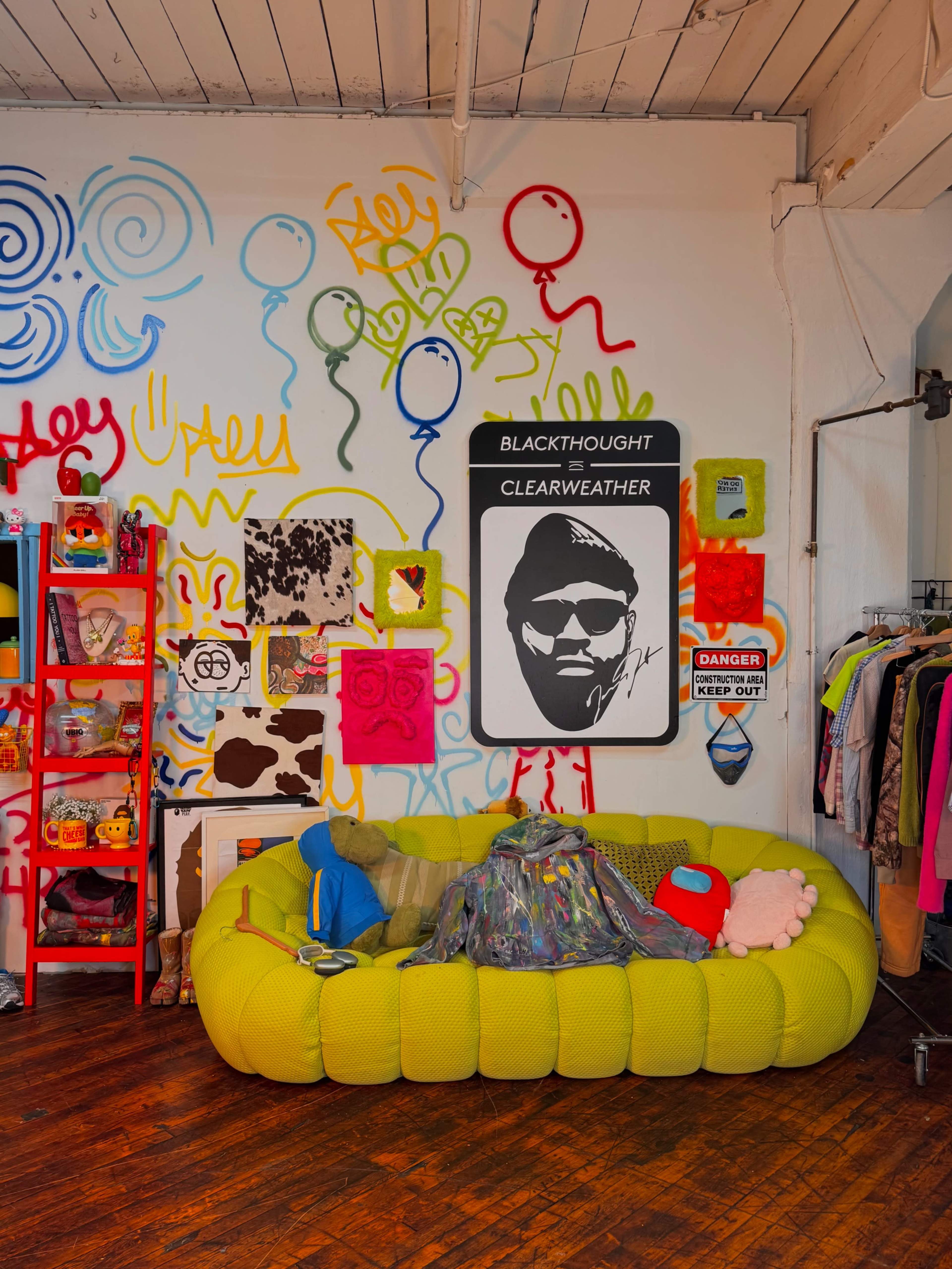 The image shows a vibrant room with a large green couch surrounded by colorful graffiti on the walls and various clothing items and accessories displayed on racks.