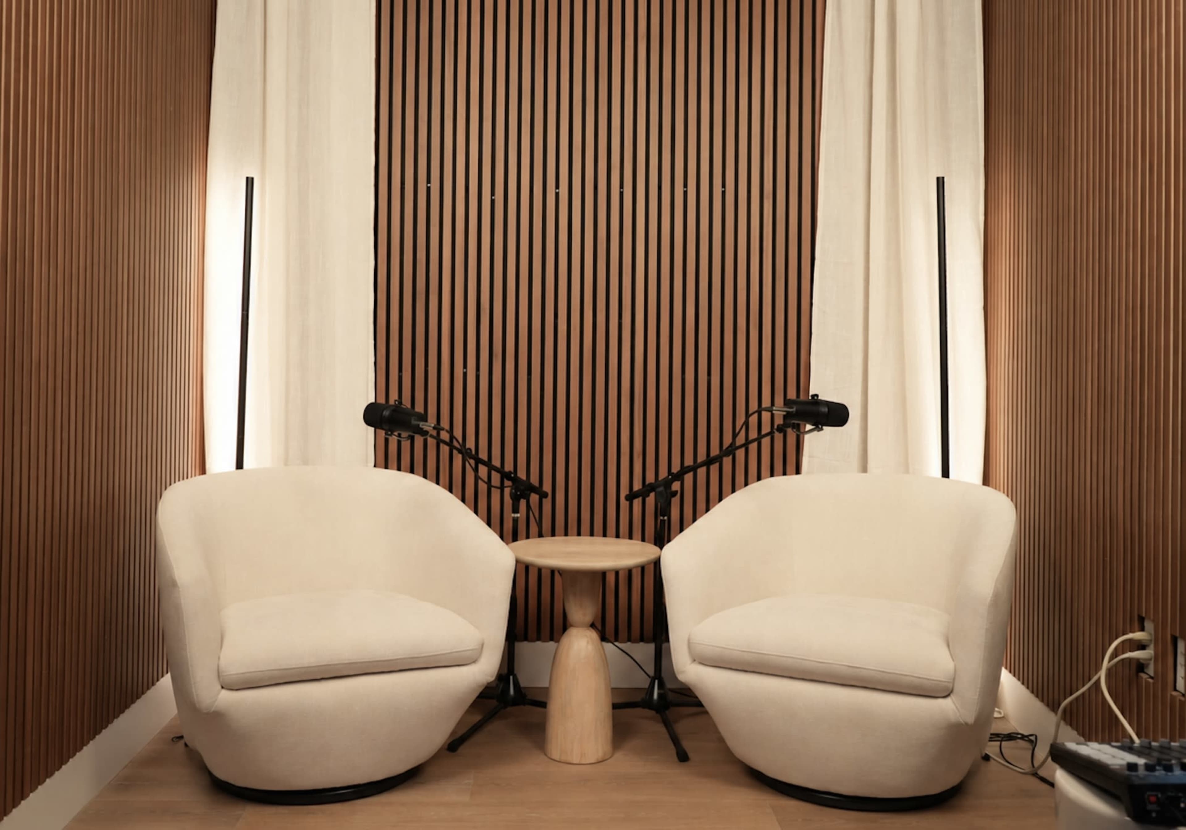 The image features two beige chairs positioned facing a small wooden table, against a backdrop of vertical wood slats and curtains, with microphones set up on stands nearby.