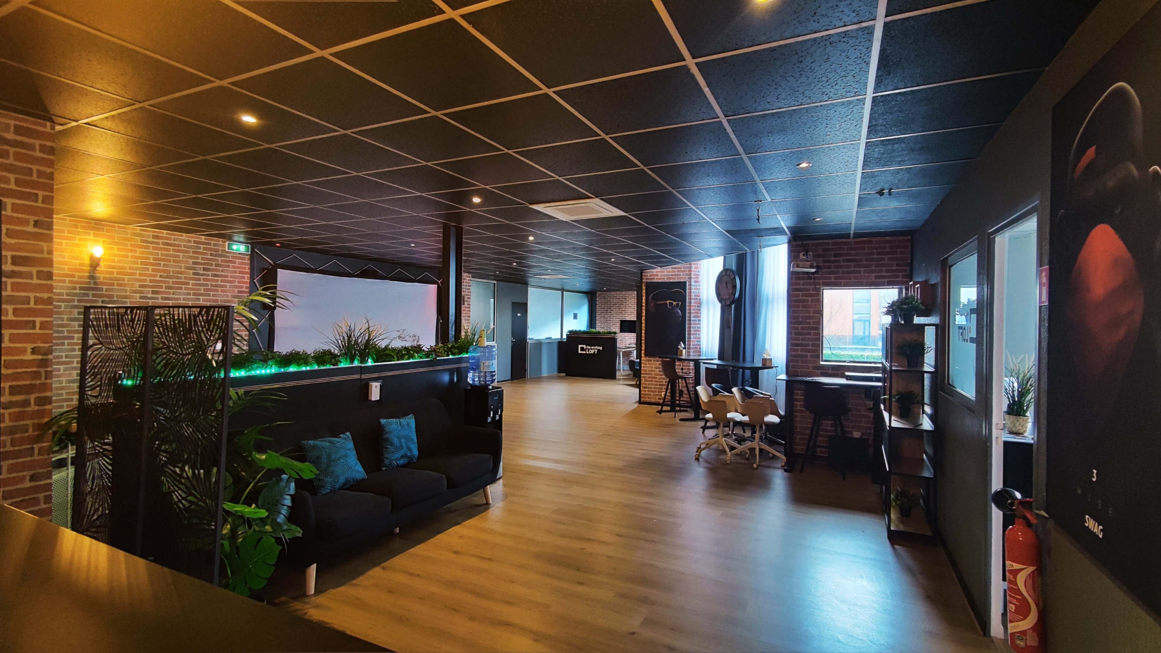 A spacious office interior featuring a lounge area with a black sofa, plants, and modern furnishings, alongside a large open workspace with a polished wooden floor.
