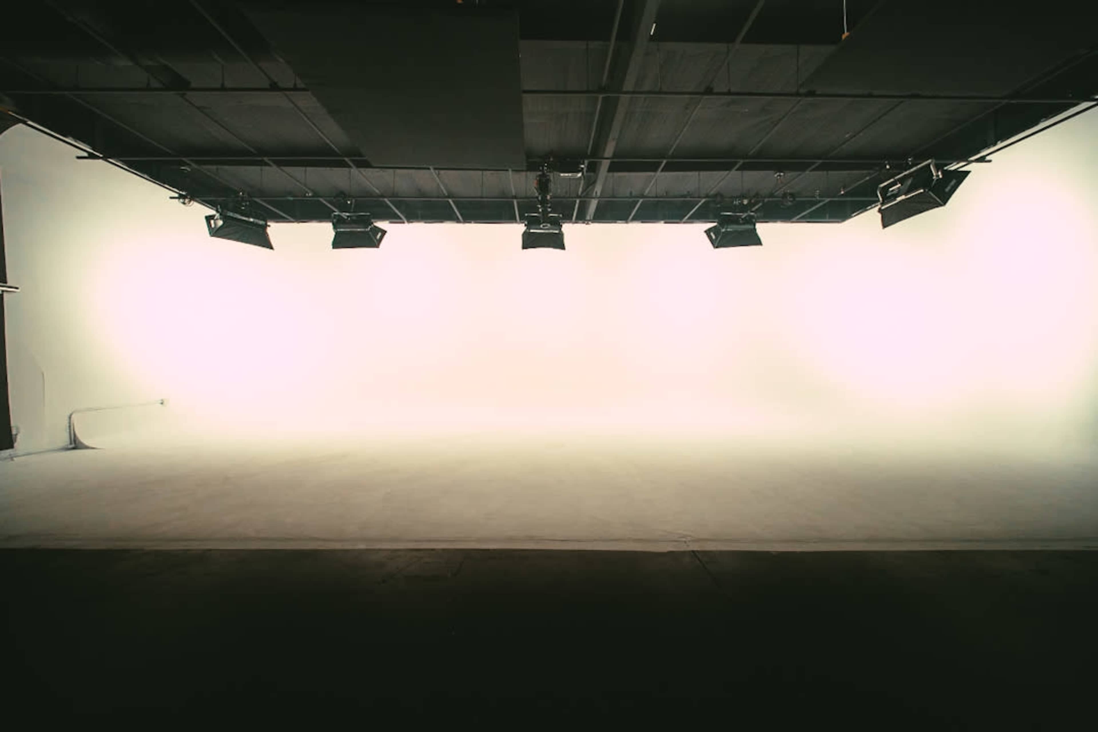 The image shows a spacious, minimally furnished studio with a blank white backdrop and overhead lighting.