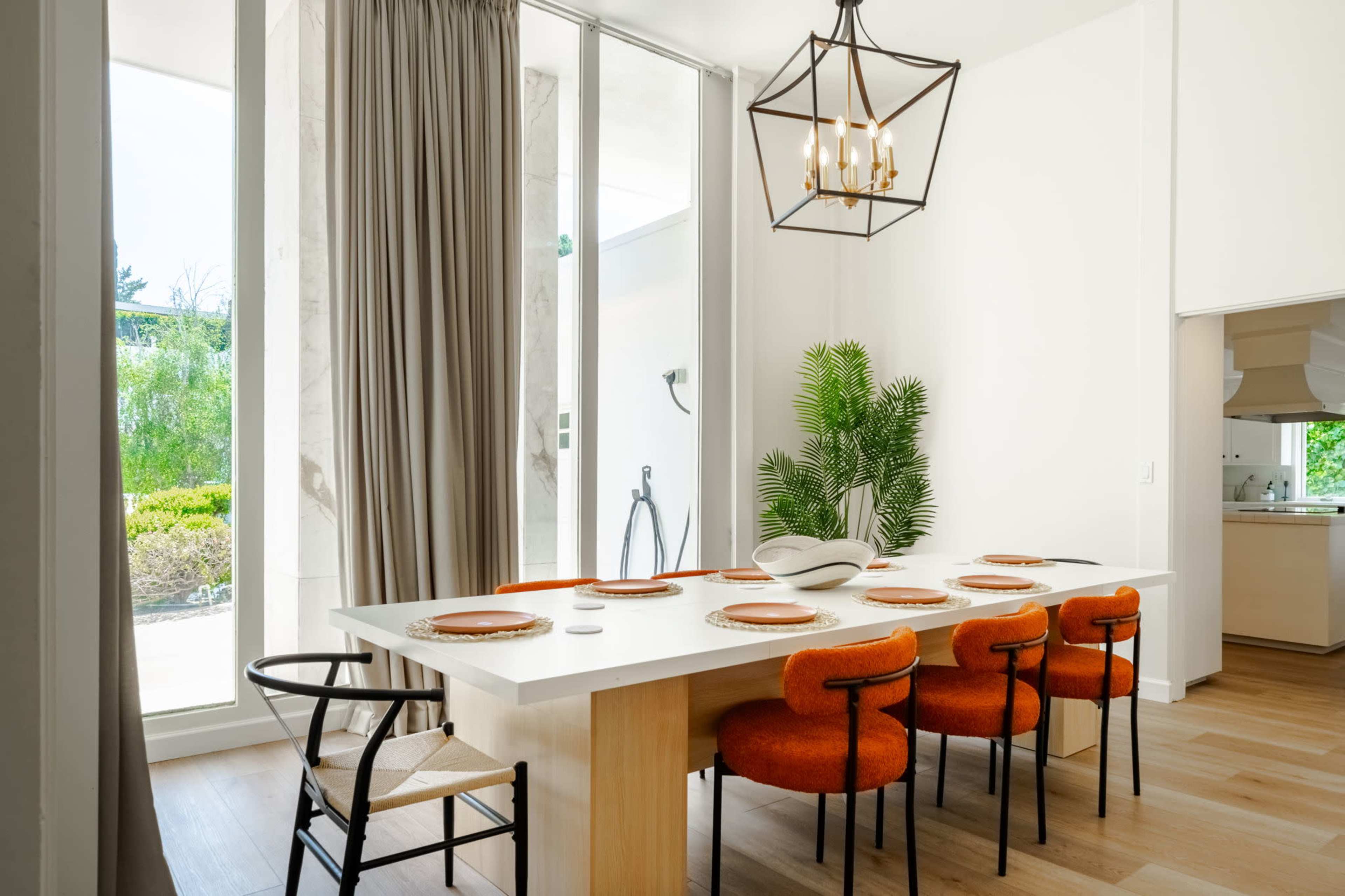 A modern dining area features a long table with orange chairs, warm wood flooring, and large windows allowing natural light.