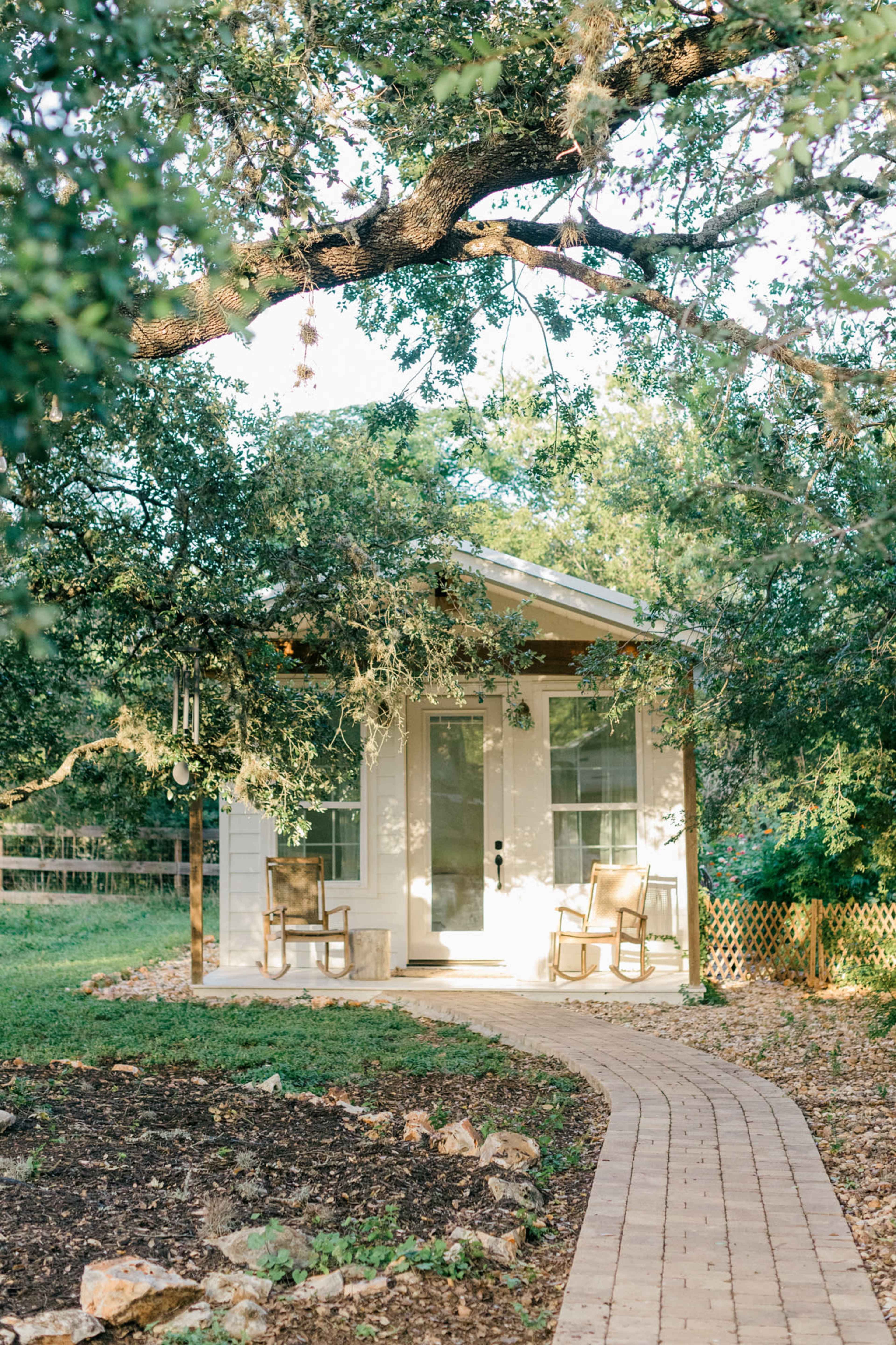 A small white cottage with two chairs on the porch is surrounded by trees and a winding stone path.