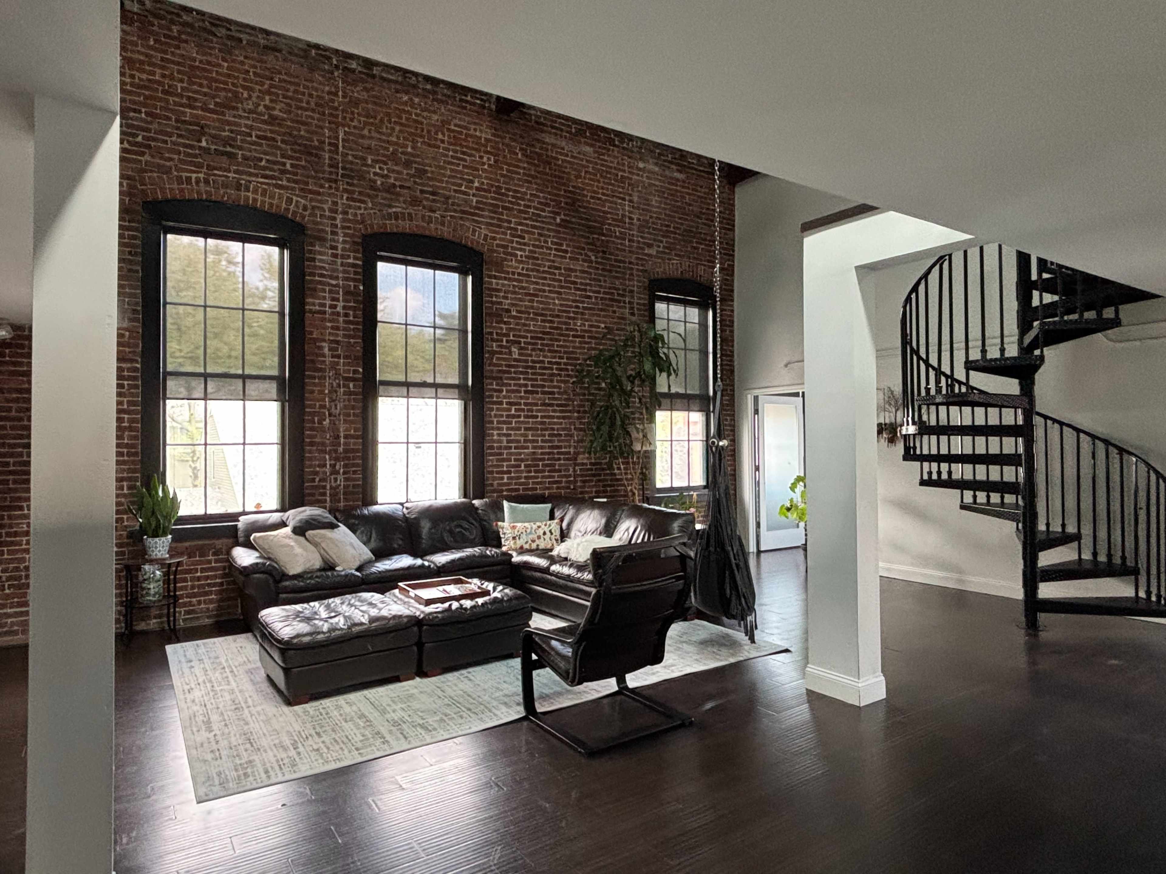 The image shows a spacious living room with exposed brick walls, large windows, and a dark spiral staircase leading to an upper level.