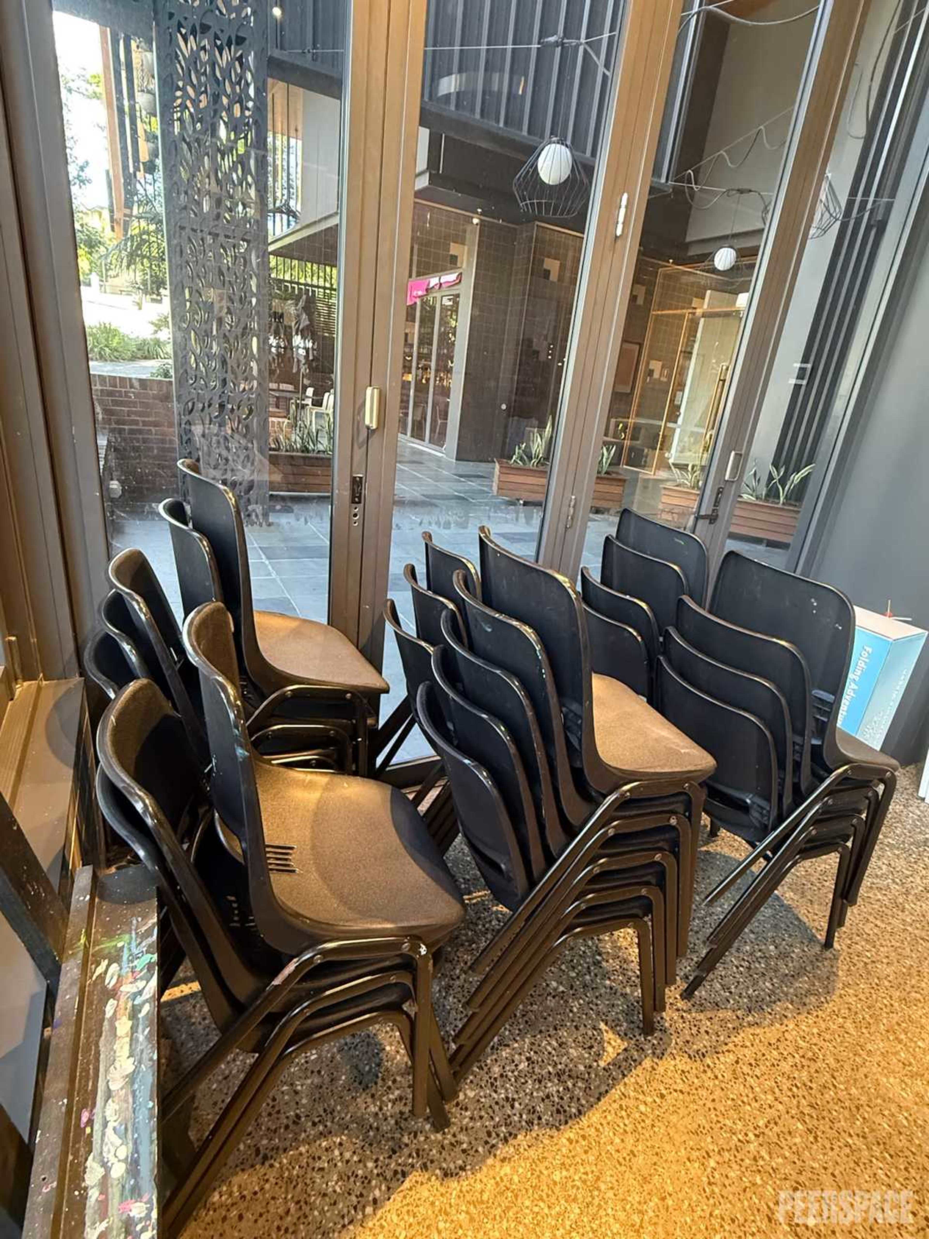 A stack of black chairs is piled neatly against a large window in a modern indoor space.