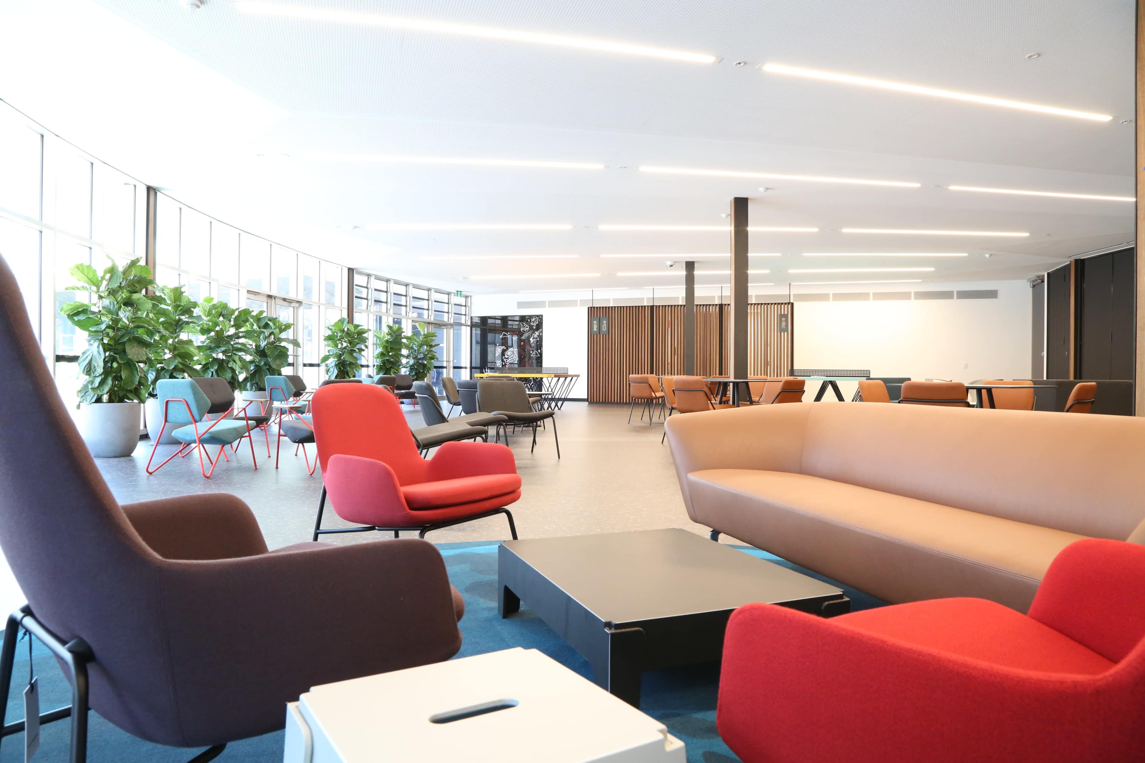 A modern lounge area with various chairs and a low table arranged around a carpet, featuring large windows and greenery in the background.