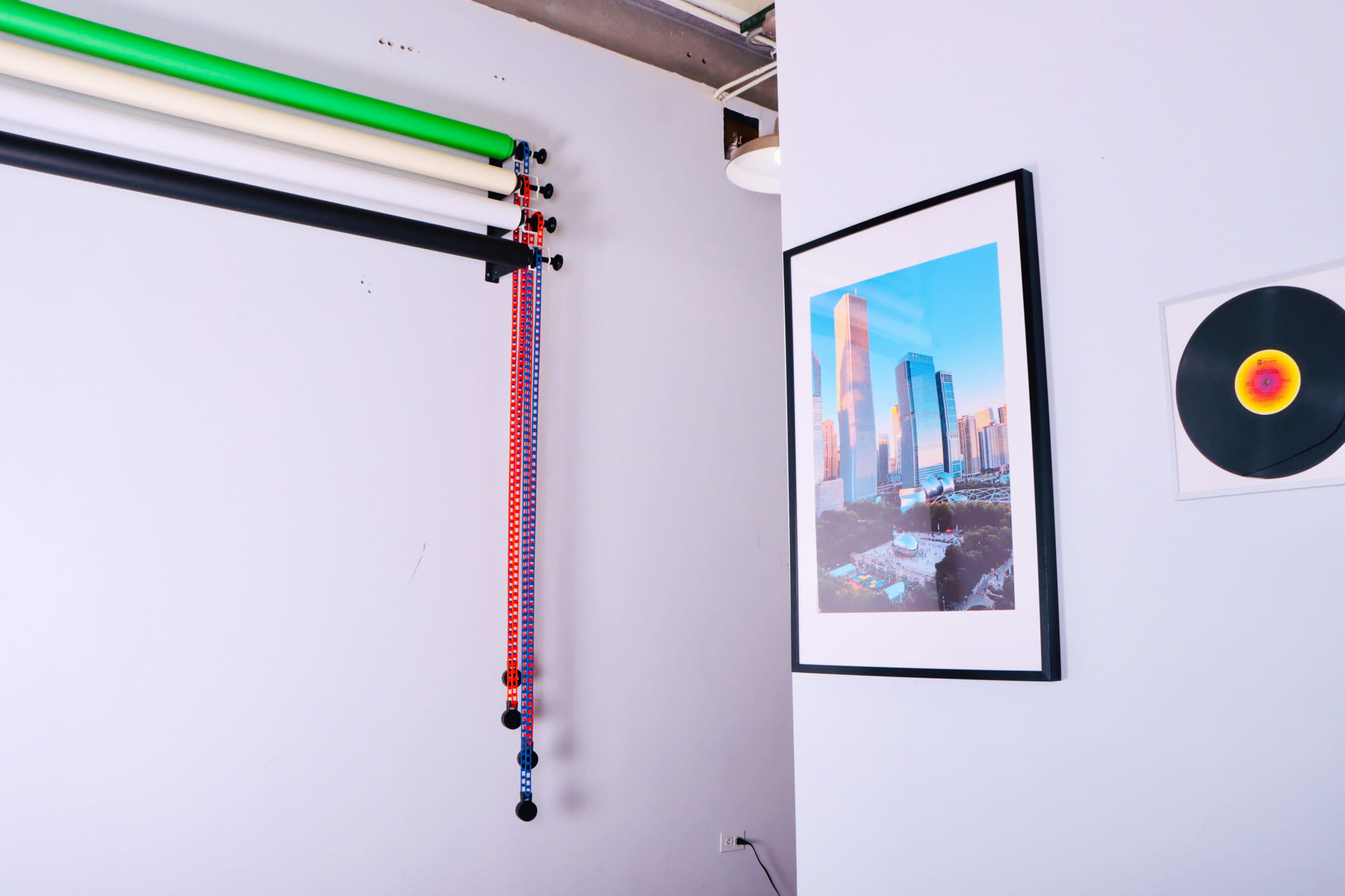 A corner of a room with colored backdrop rolls mounted on the wall, a framed photograph of a city skyline, and a vinyl record displayed on another wall.