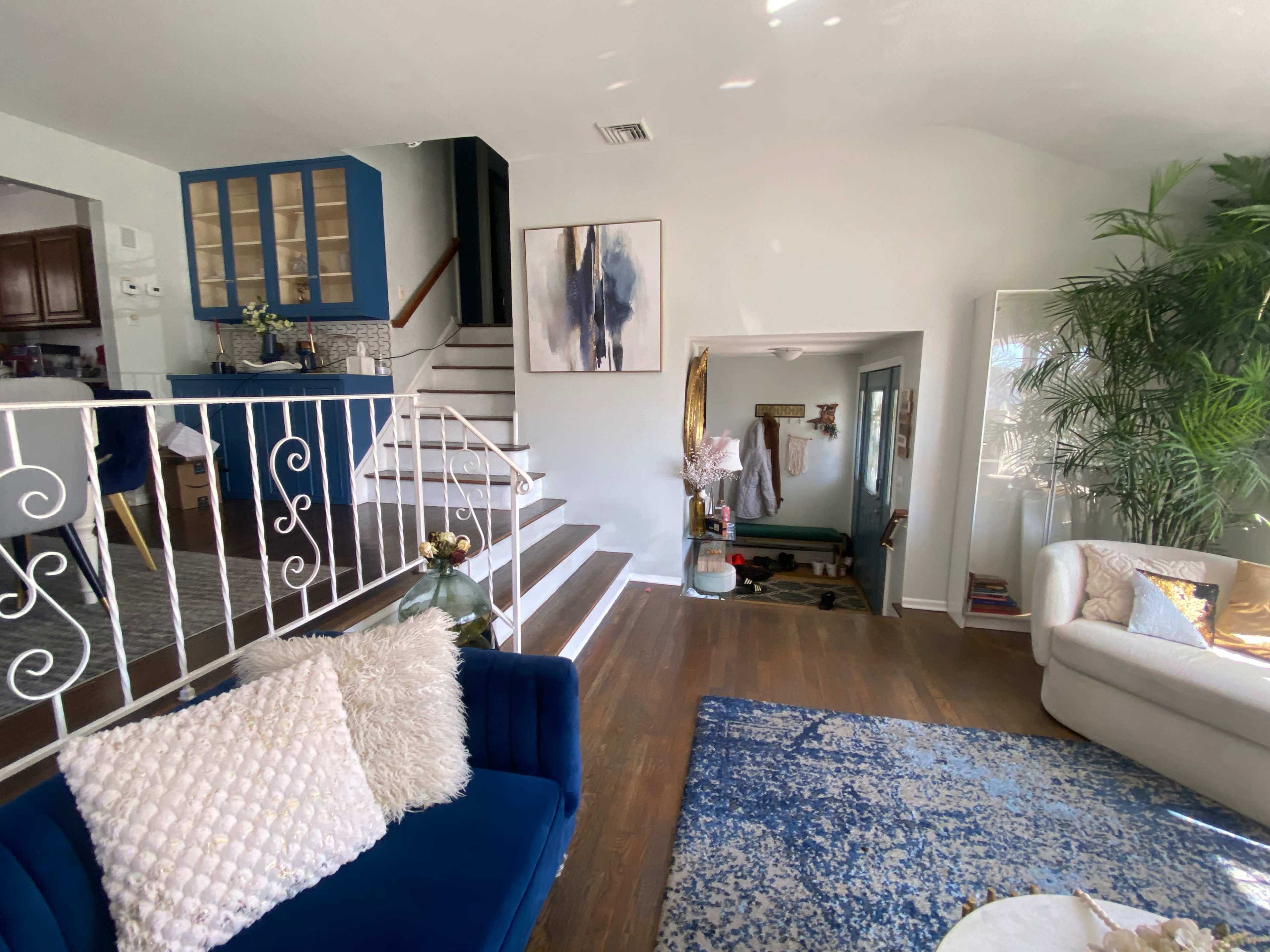 The image shows a well-lit living space featuring a blue couch, a large area rug, and a staircase leading to an upper level, with a glimpse of a kitchen in the background.