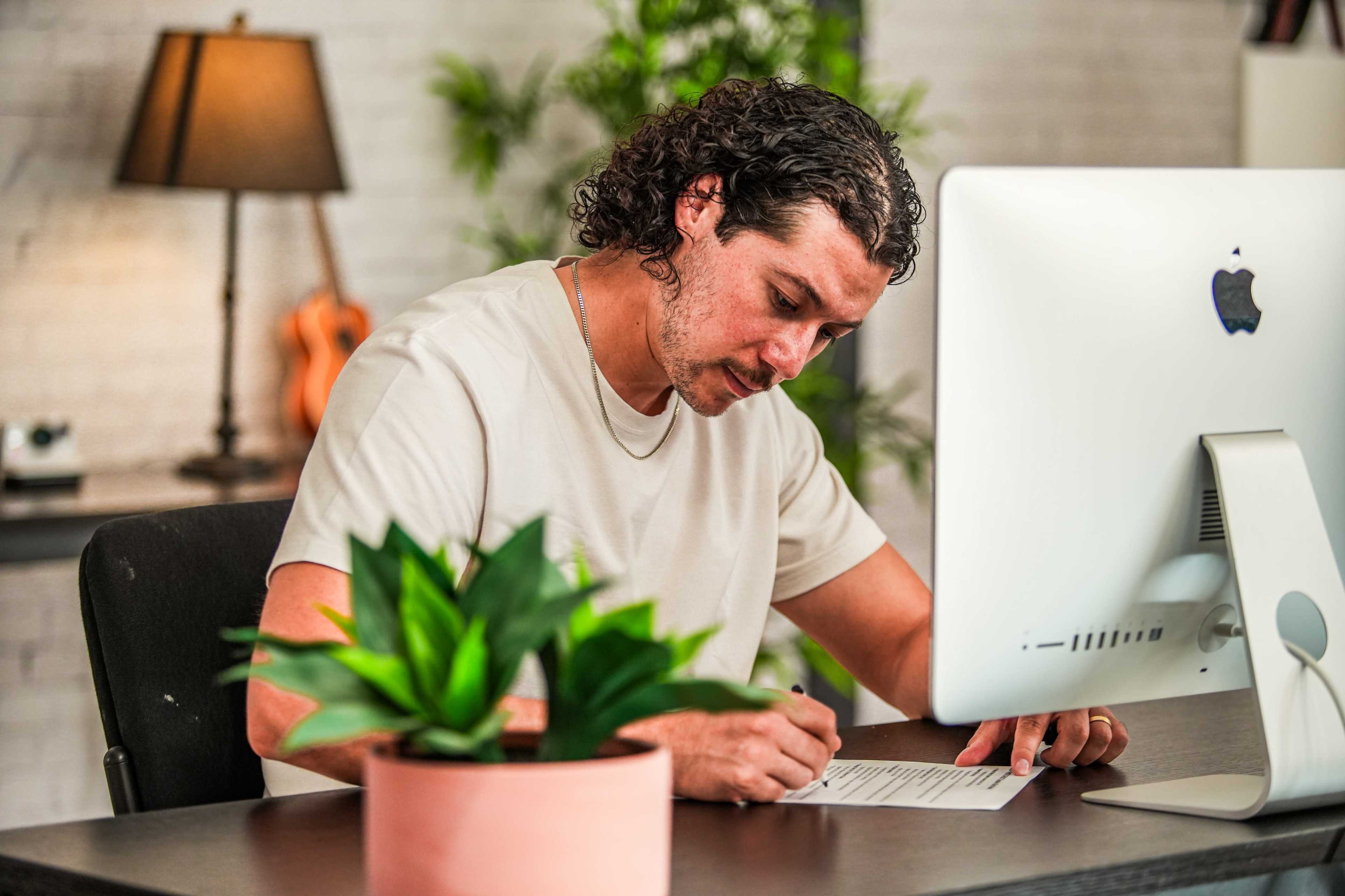 A person sits at a desk, writing on paper while facing a computer monitor and a potted plant.