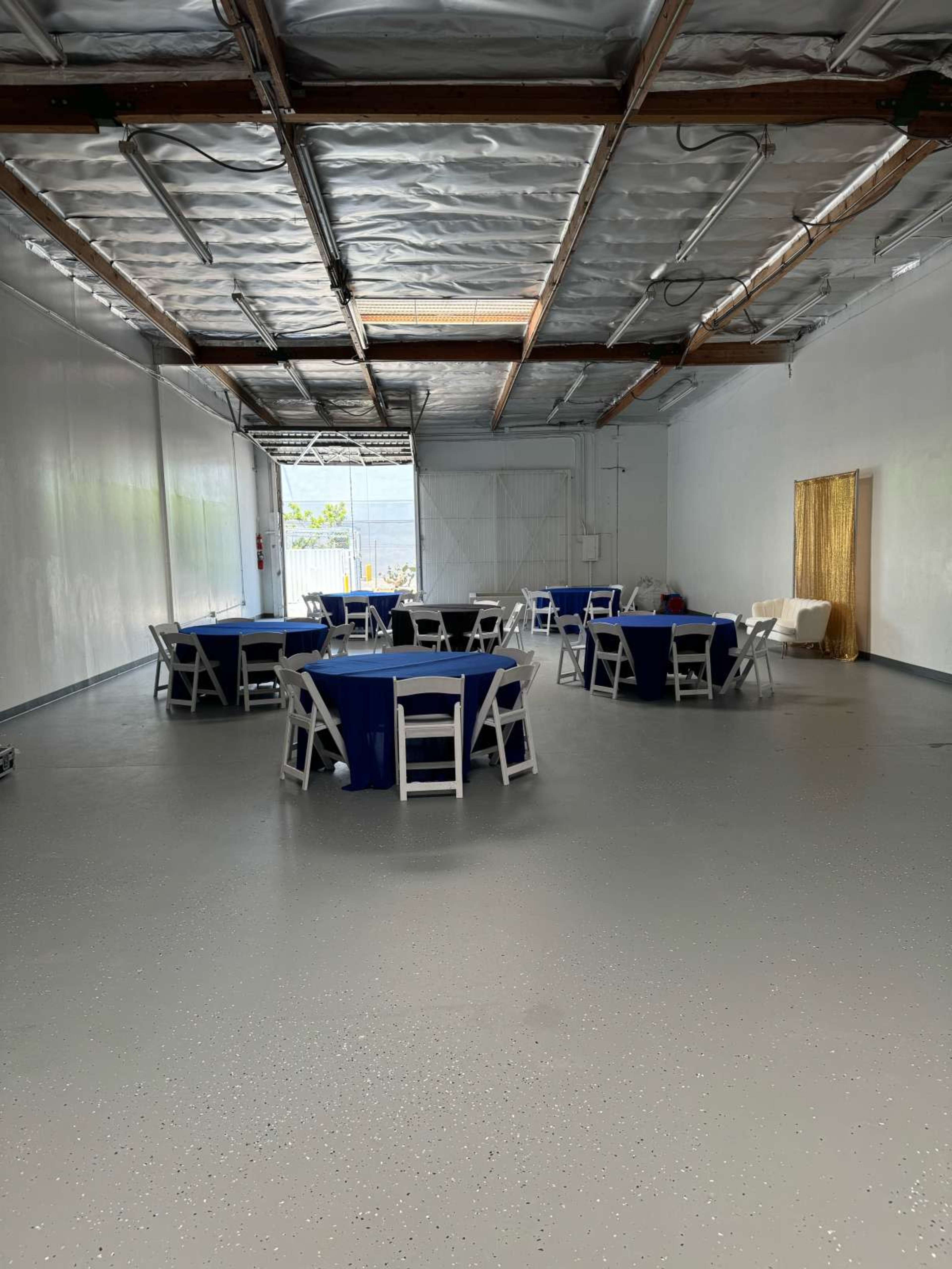An empty indoor space with several round tables covered in blue tablecloths and white chairs arranged around them.