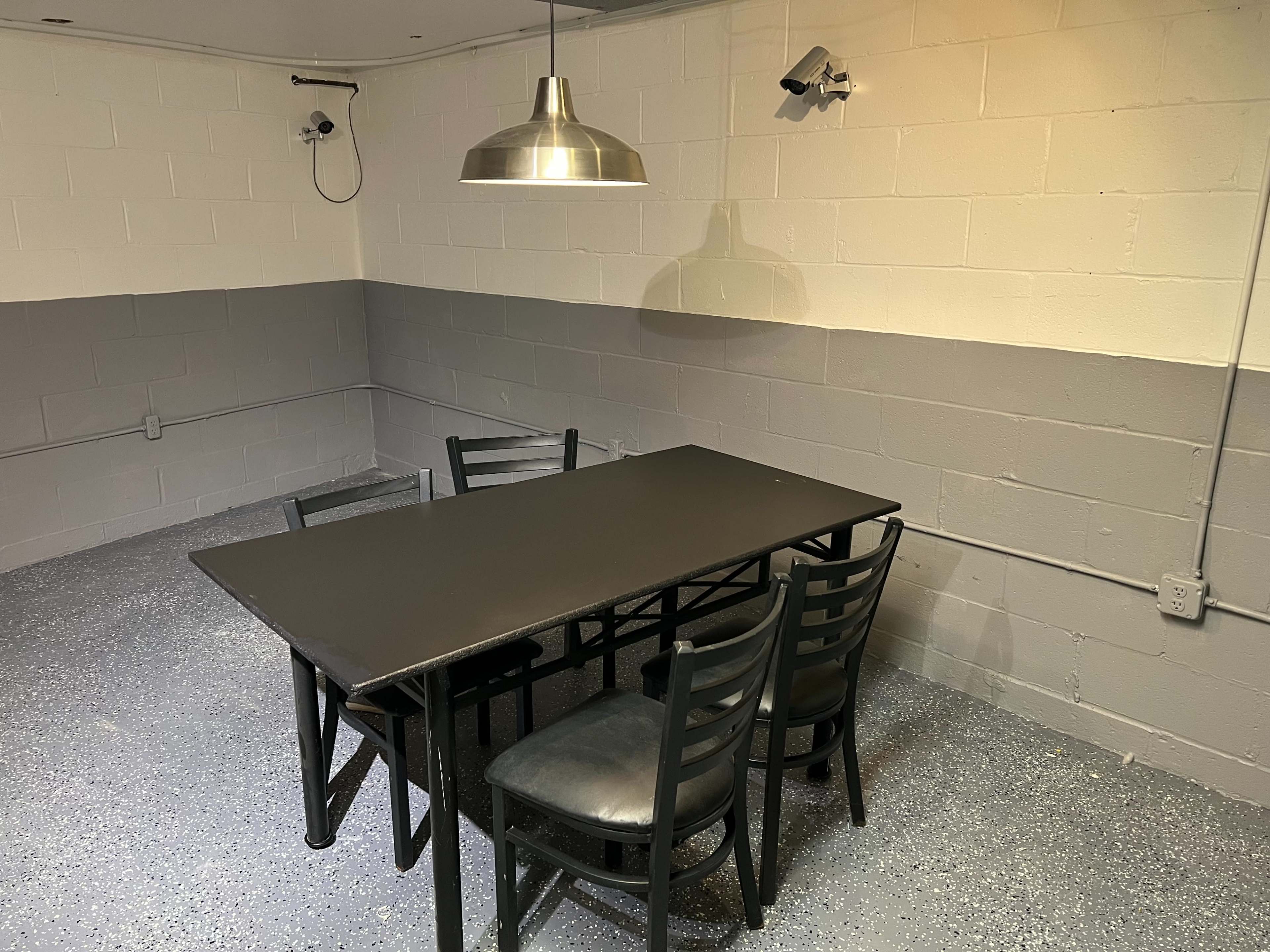 A black table and six chairs are positioned in a sparsely furnished room with gray-painted walls and ceiling-mounted cameras.