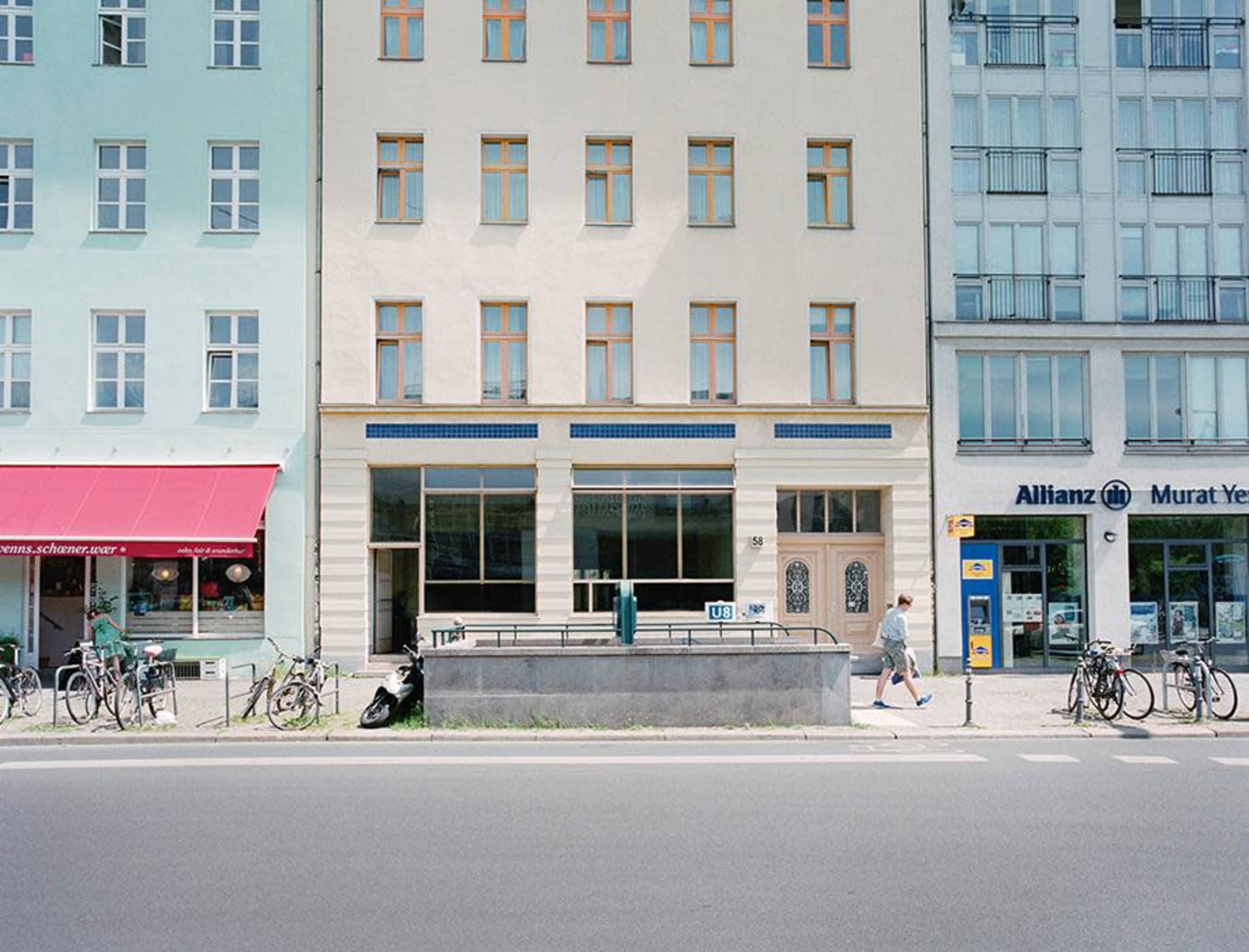 The image shows a building facade with multiple windows, flanked by shops and bicycles parked along a sidewalk, and a street scene with a person walking by.