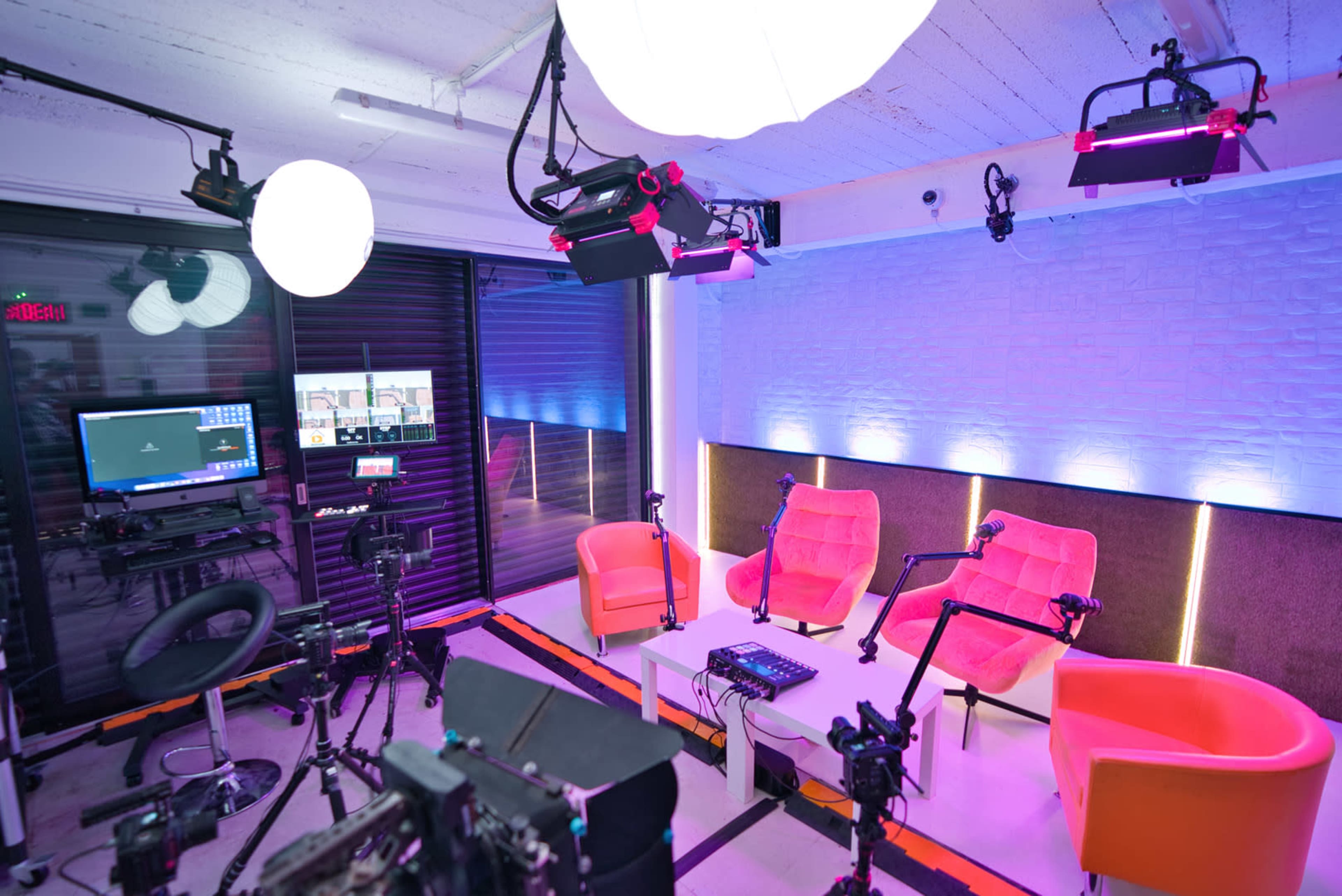 A modern studio setup with pink chairs, various lighting equipment, and multiple cameras arranged around a central table.