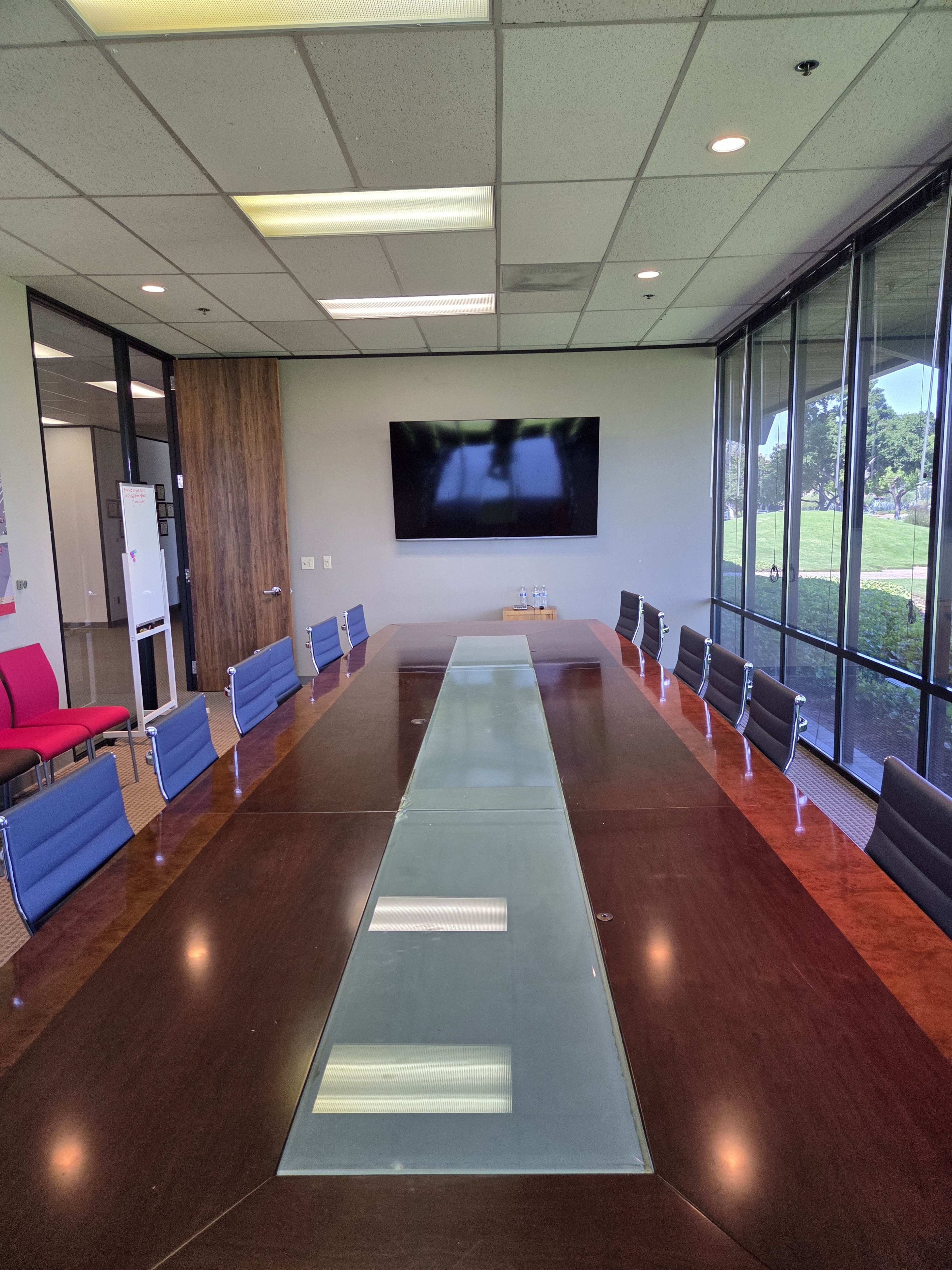 A large conference room features a long wooden table with a glass center, surrounded by several blue chairs, and glass walls overlooking an outdoor area.