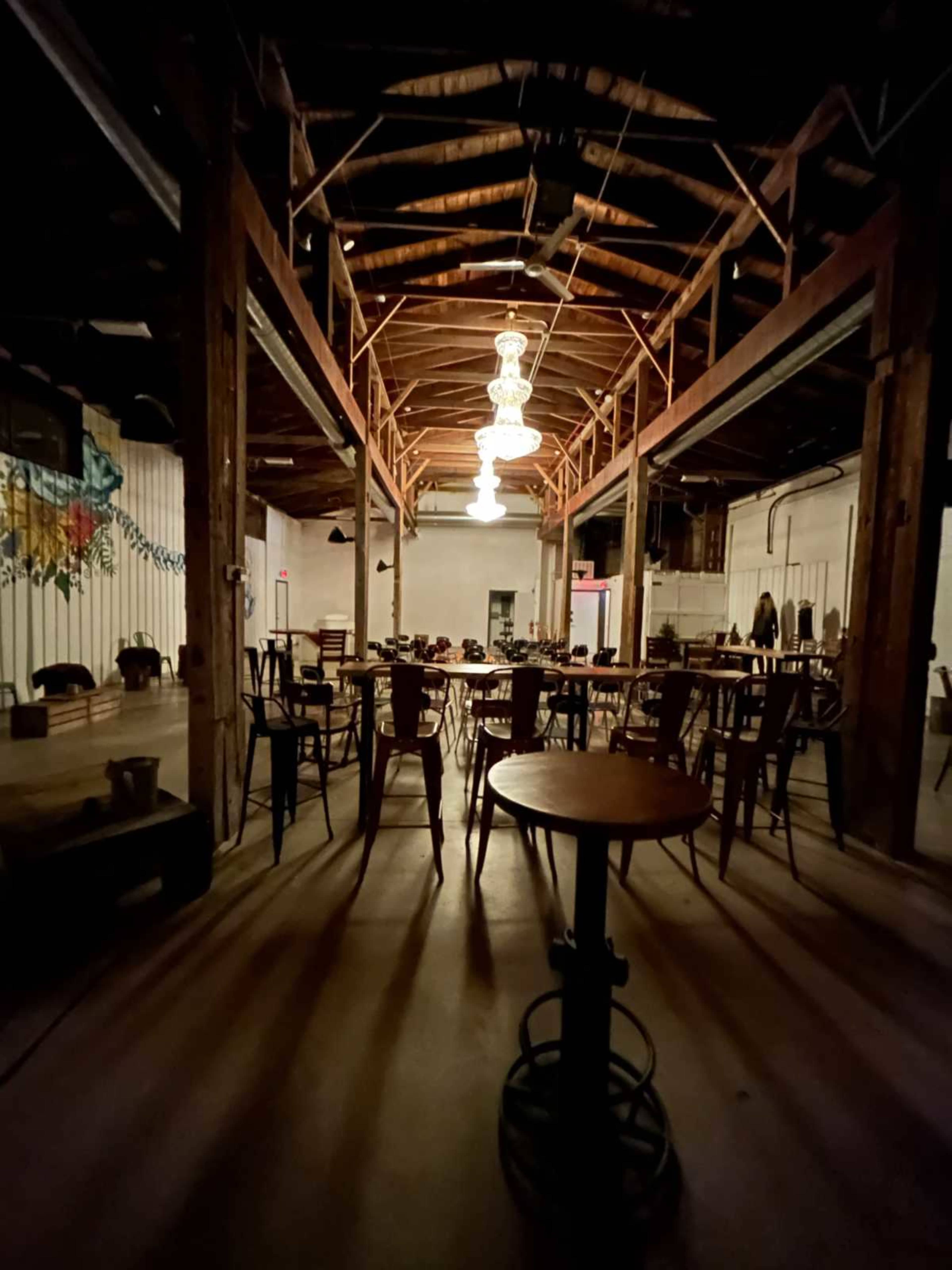 The image shows a dimly lit interior of a restaurant or event space with wooden beams and numerous tables and chairs arranged throughout the room.