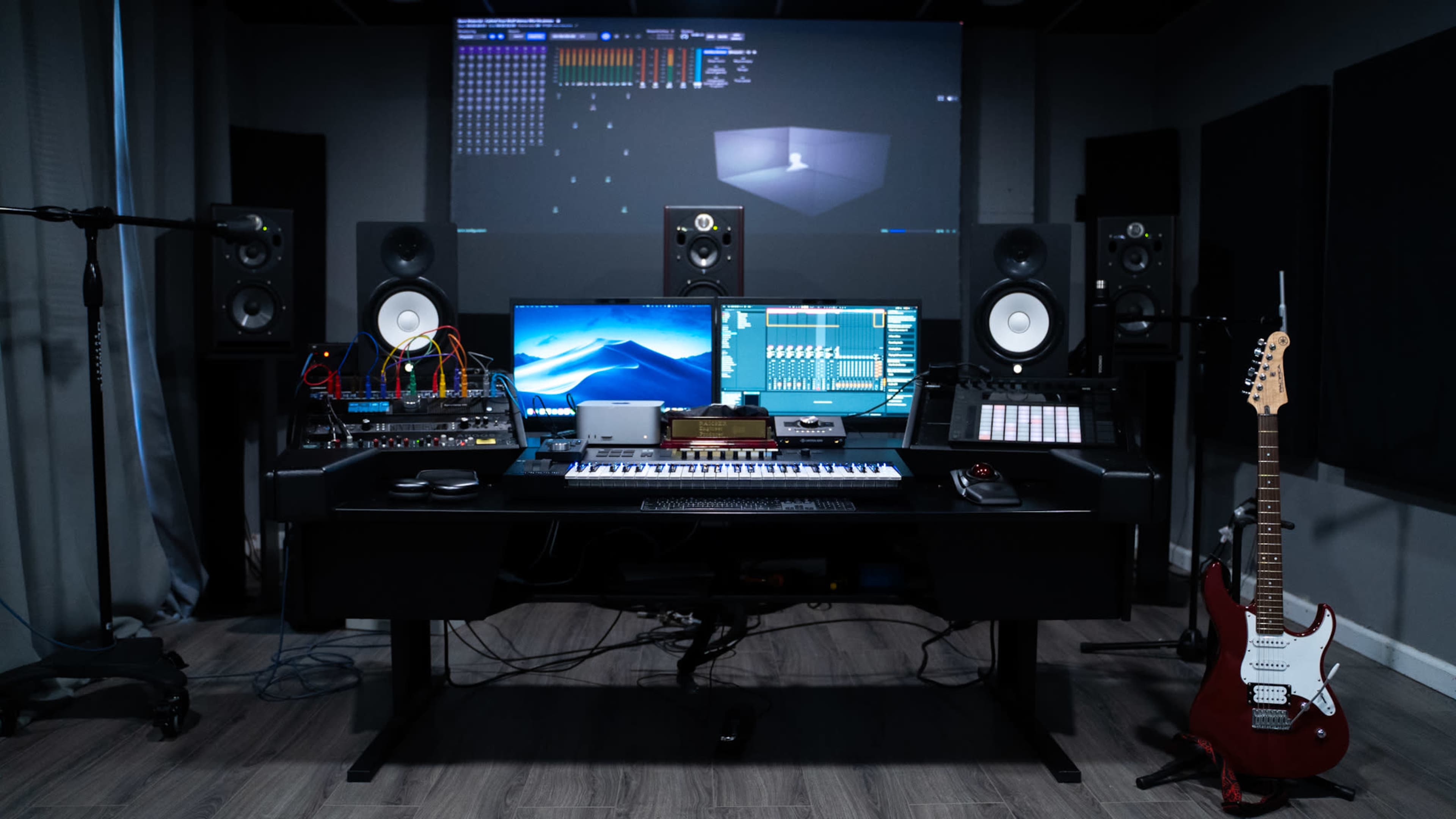 A music studio setup with a black desk, two computer monitors, audio equipment, speakers, and an electric guitar on the right.