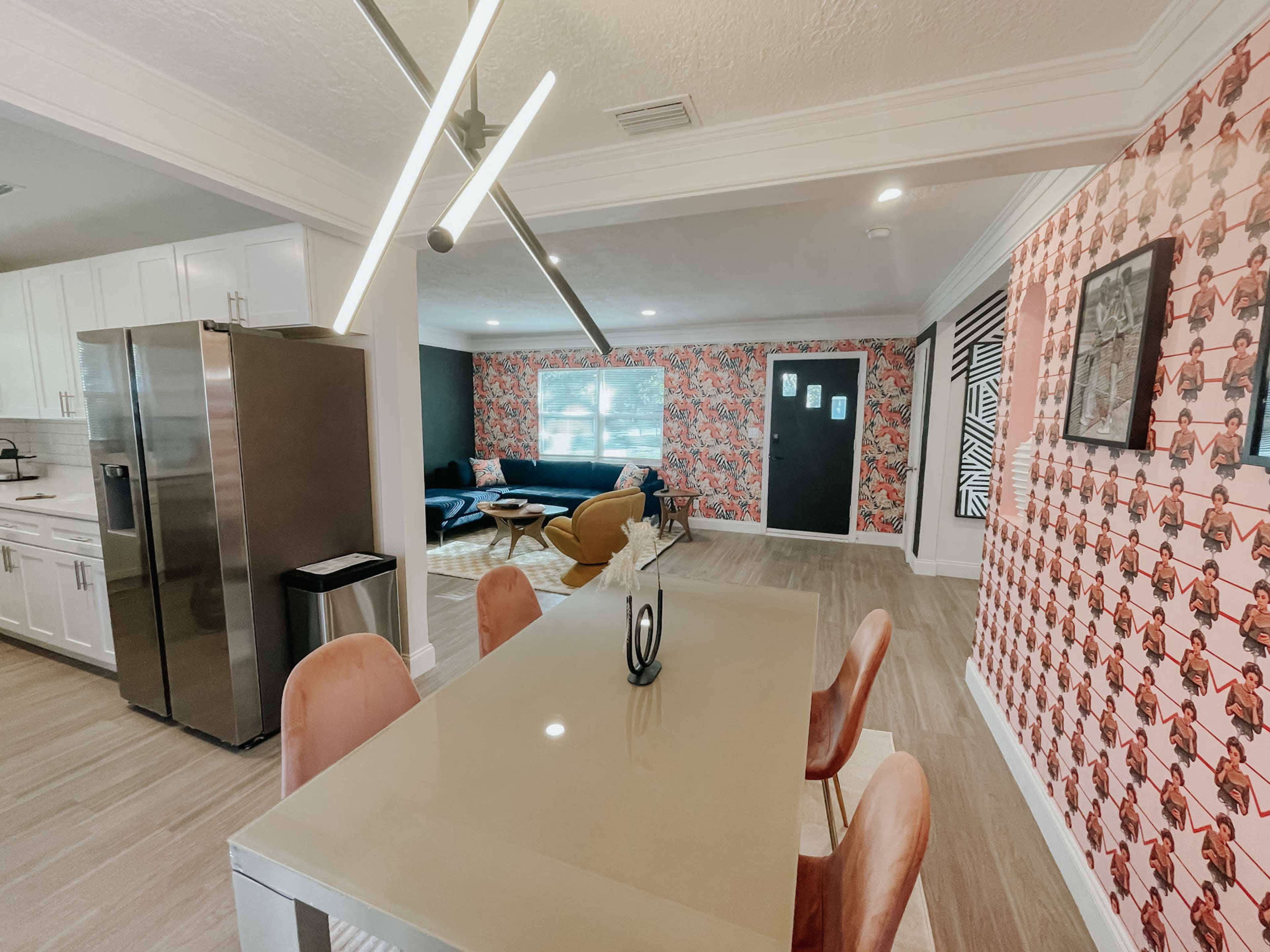 The image shows a modern dining area with a sleek table and chairs, adjacent to a living space featuring colorful wallpaper and contemporary furniture.
