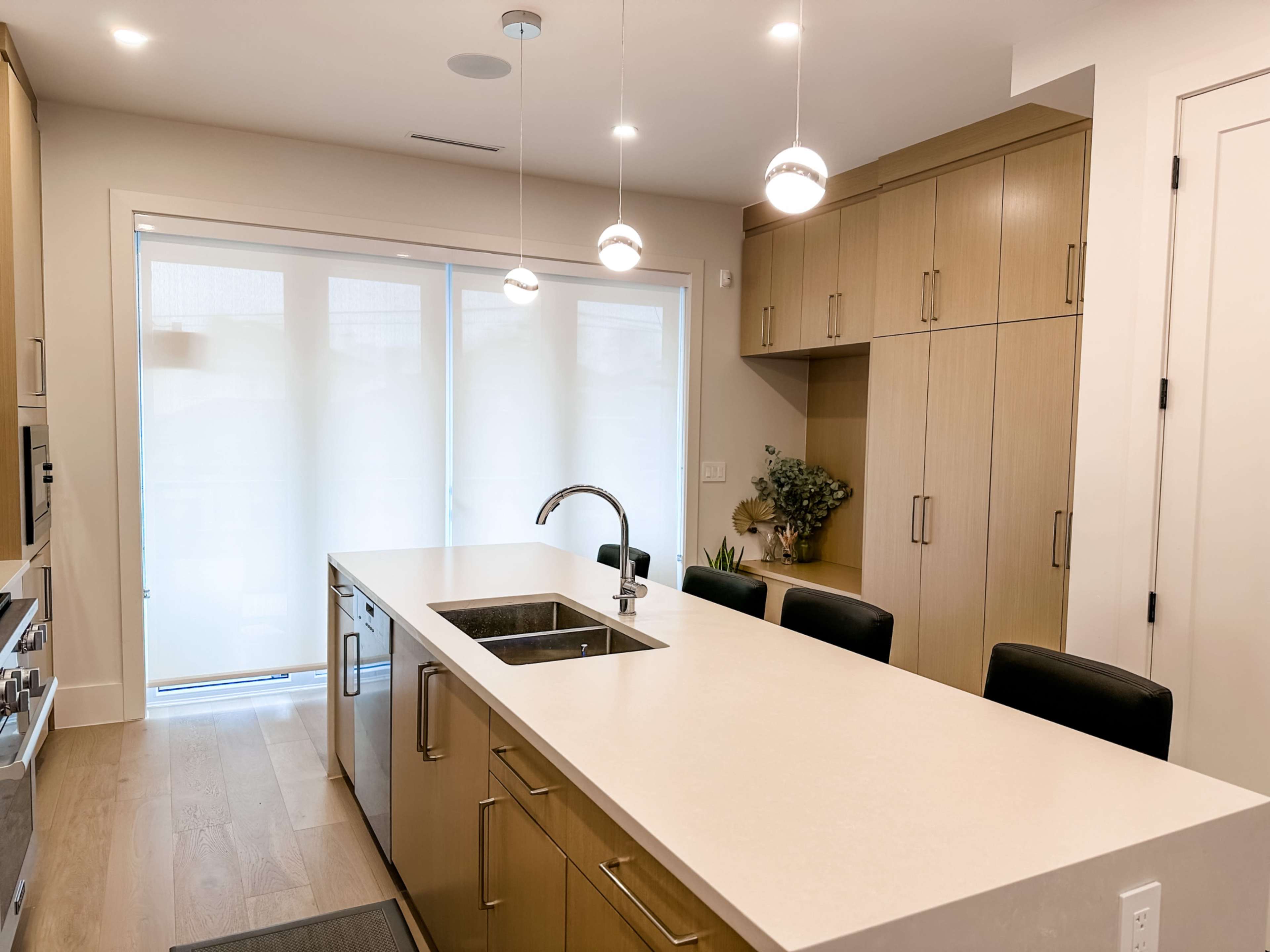 A modern kitchen features a central island with a sink and seating, light wood cabinetry, and large windows covered with sheer blinds.