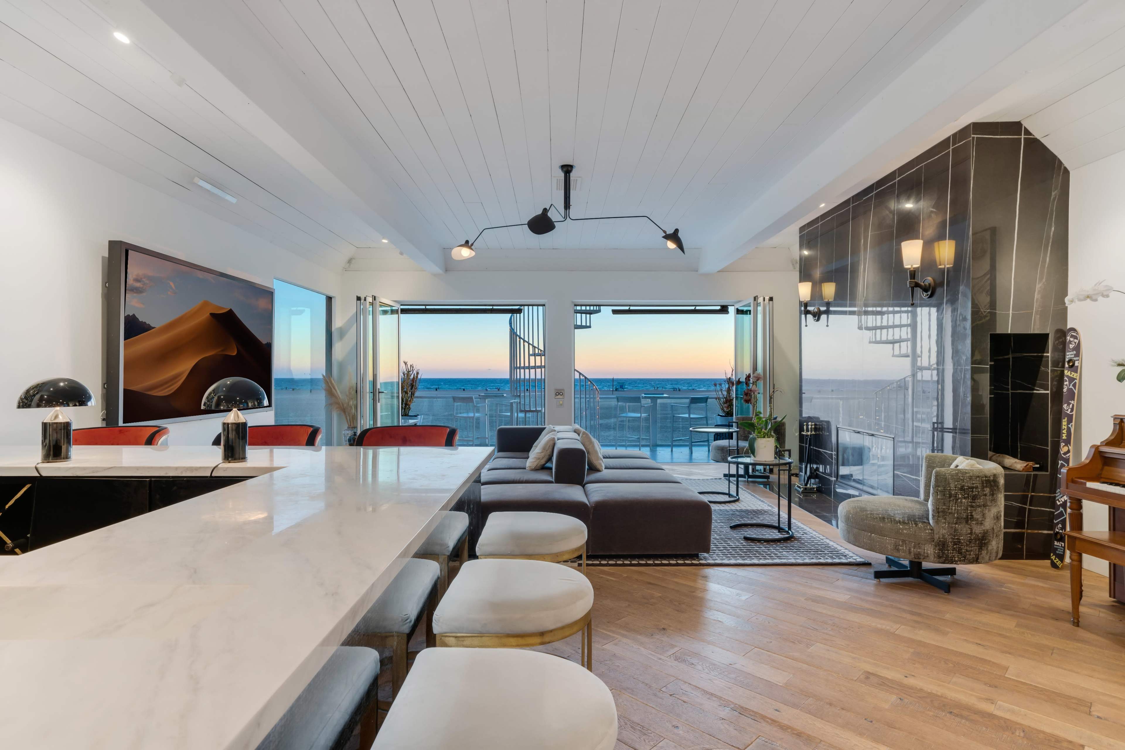 A modern living area with a large marble dining table, a sectional sofa, and expansive windows showcasing a sunset view.