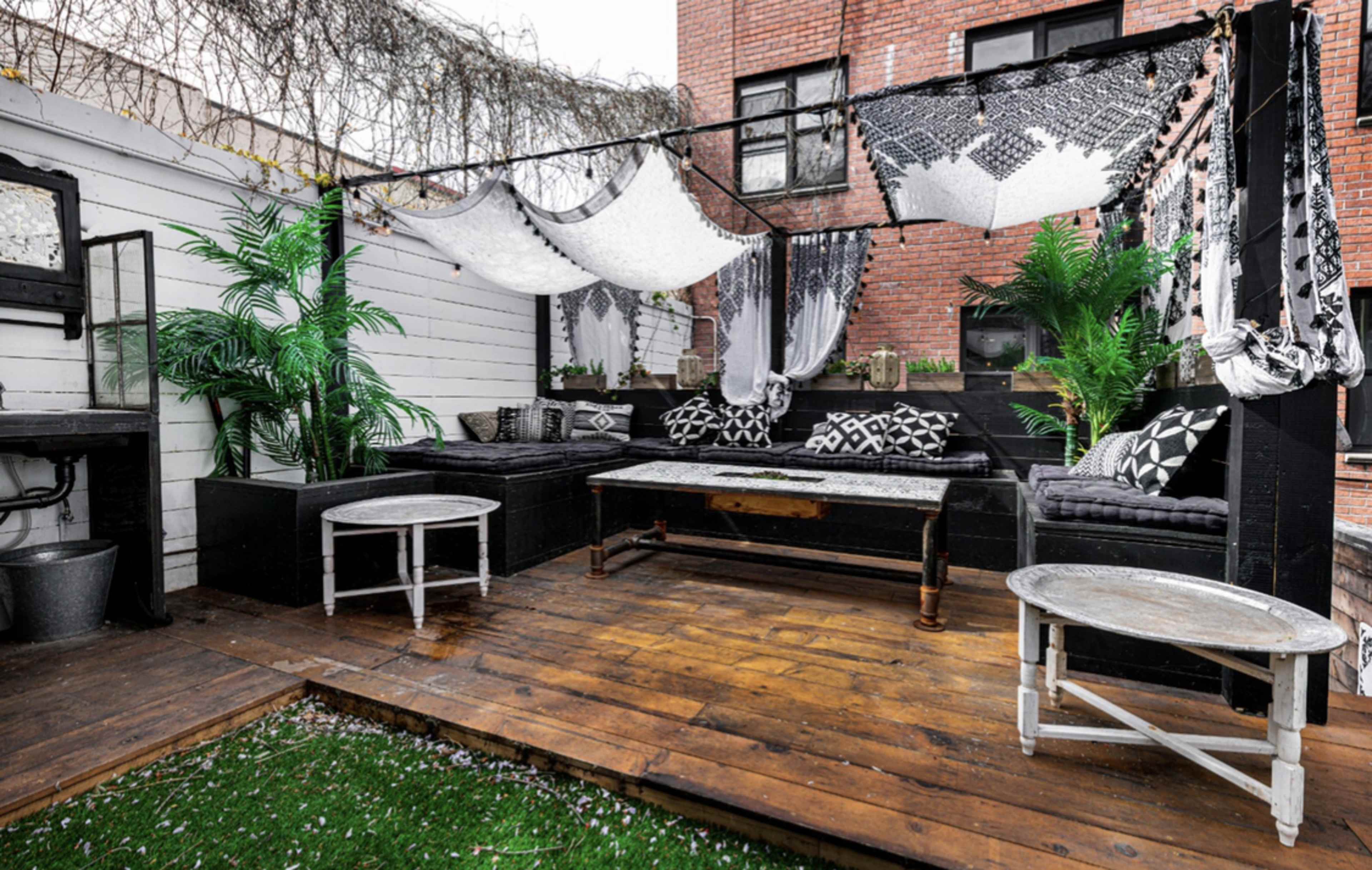 A patio features black and white seating, decorative textiles, potted plants, and wooden flooring.