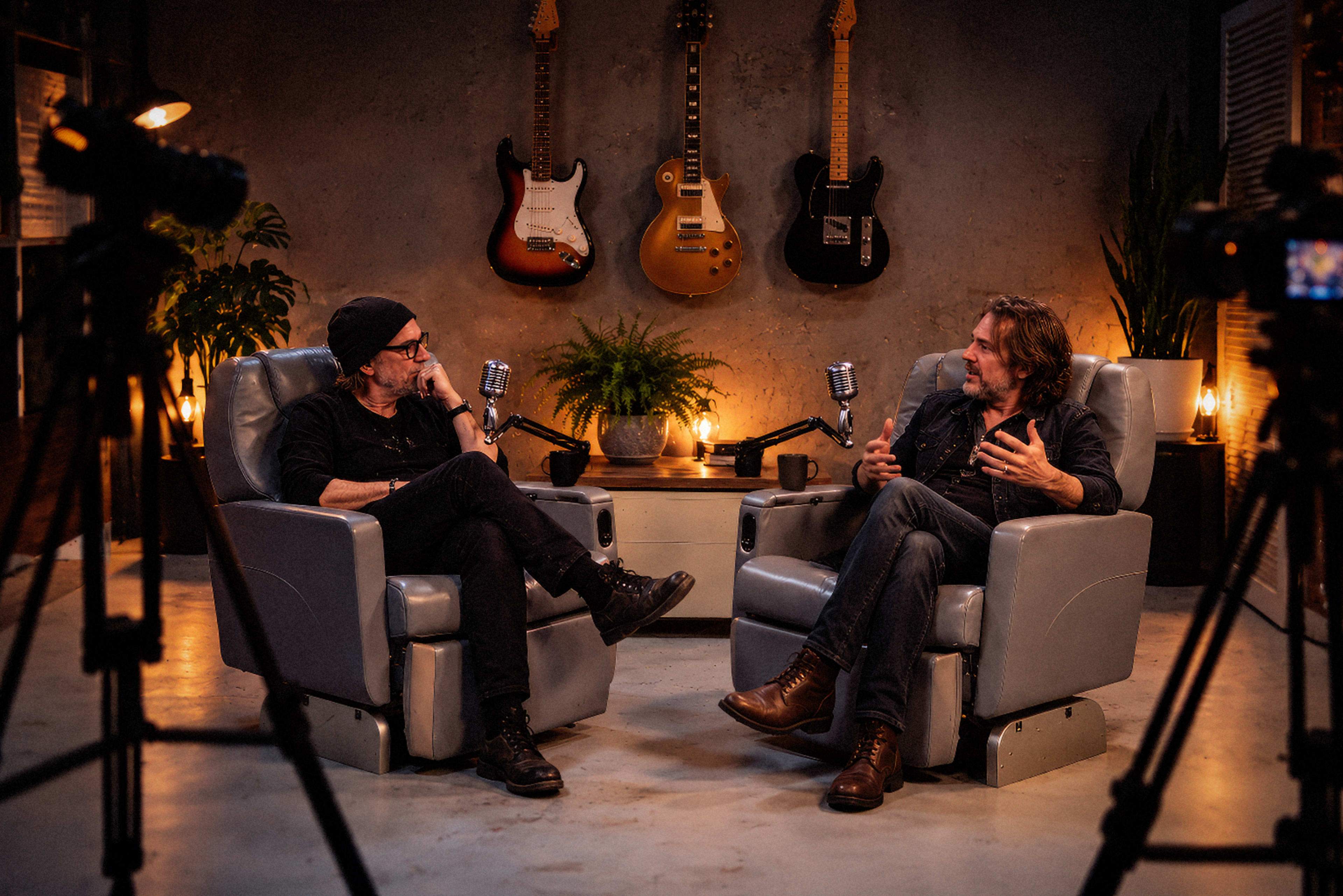 Two men are seated in gray armchairs facing each other in a well-lit room decorated with guitars and plants, with cameras positioned nearby.