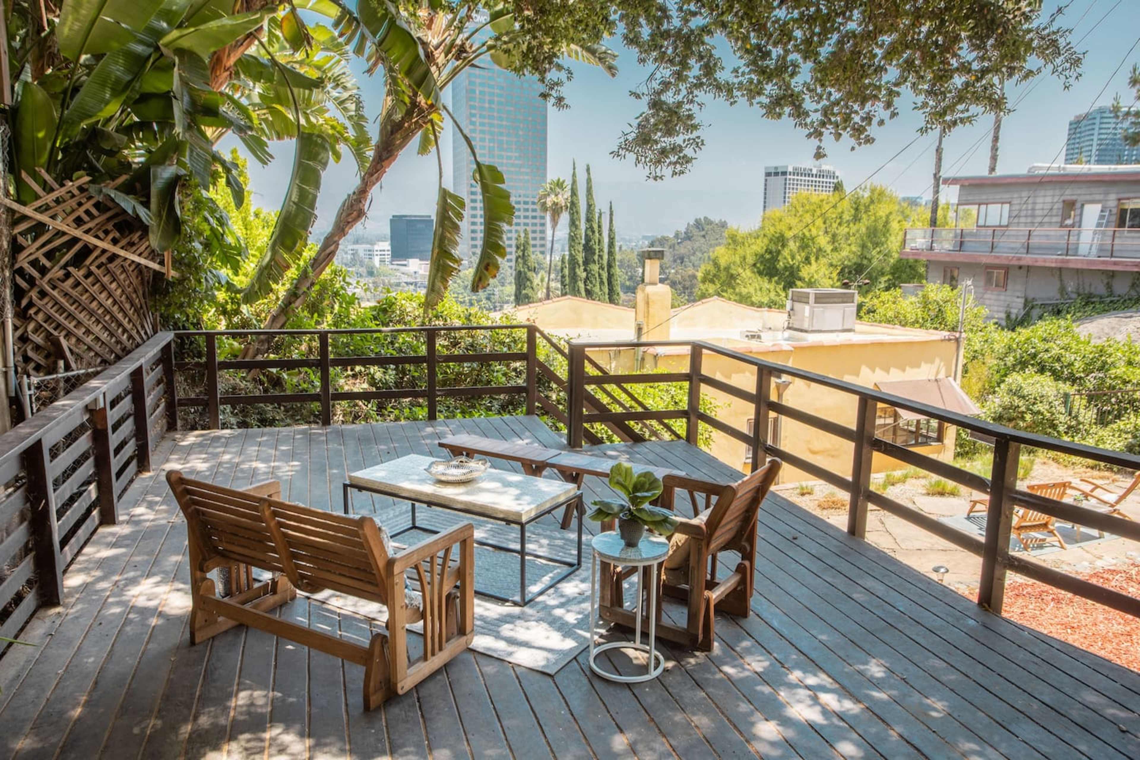 A wooden deck with a table and chairs overlooks a cityscape surrounded by greenery.