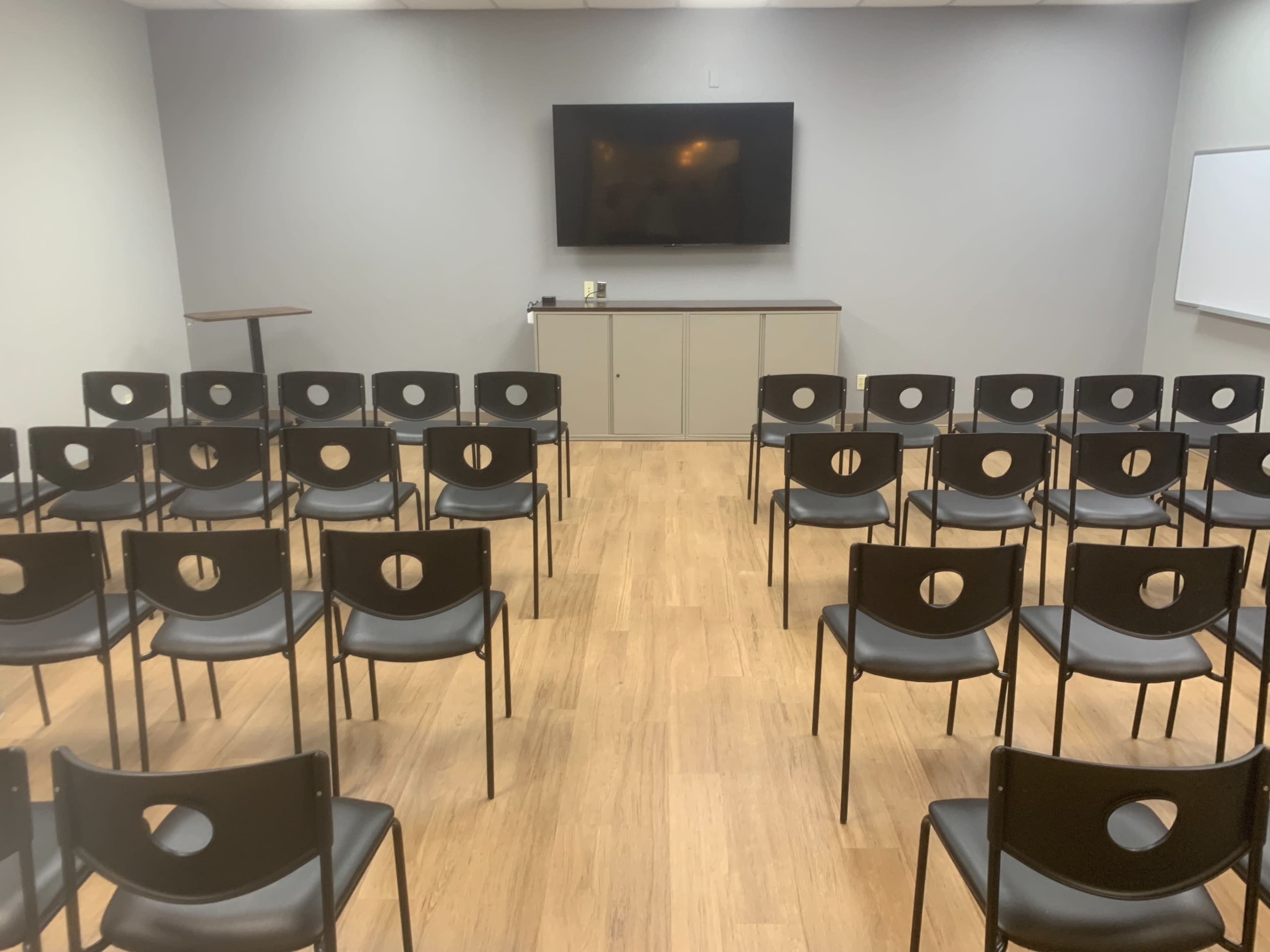 A meeting room is set up with rows of black chairs facing a wall-mounted television and a whiteboard.