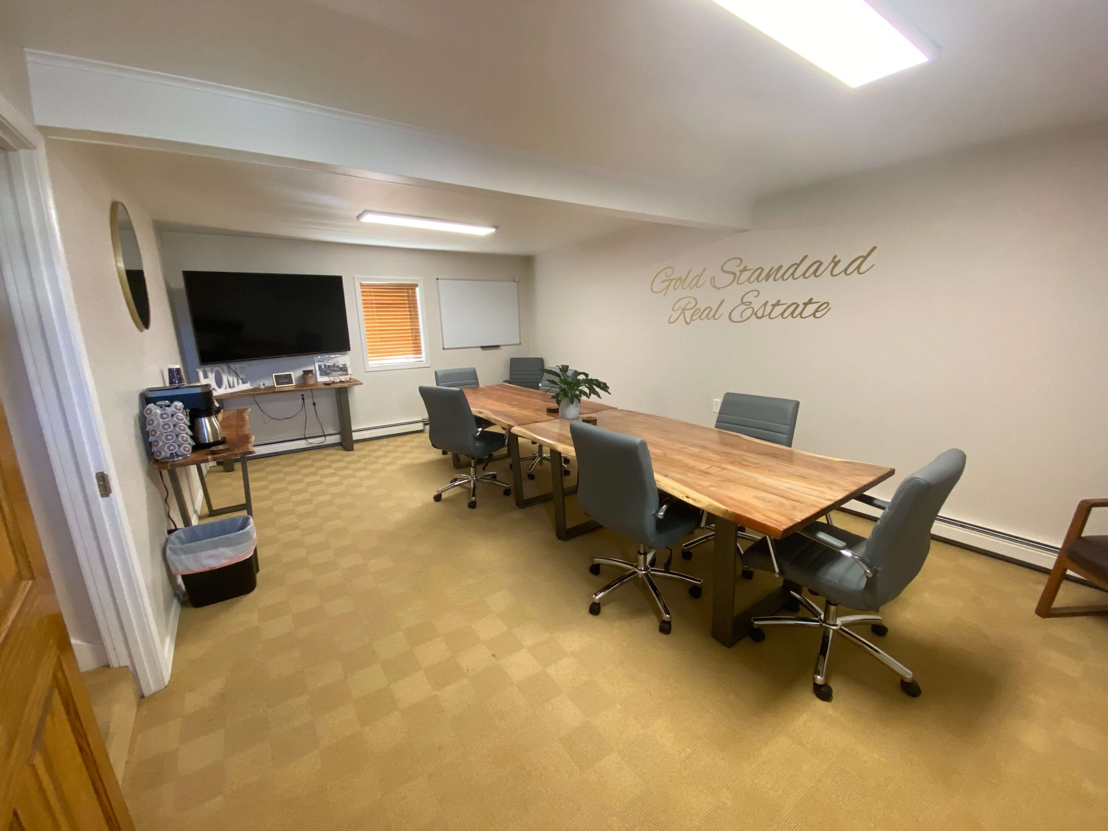 A modern conference room with a wooden table, six gray chairs, a wall-mounted TV, and a decorative sign that reads "Gold Standard Real Estate."
