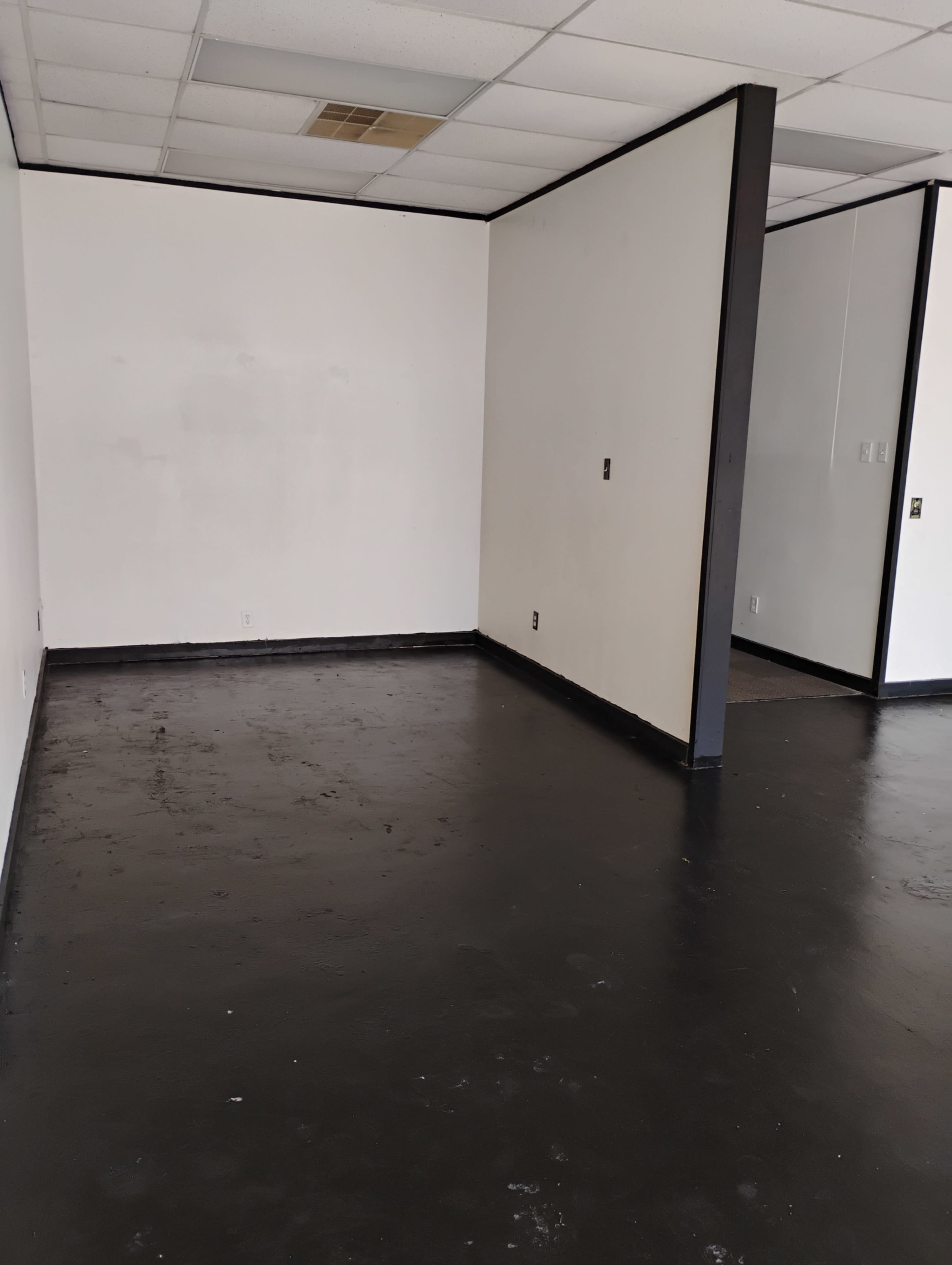 The image shows an empty room with black flooring and white walls, featuring a partially partitioned section on the right.