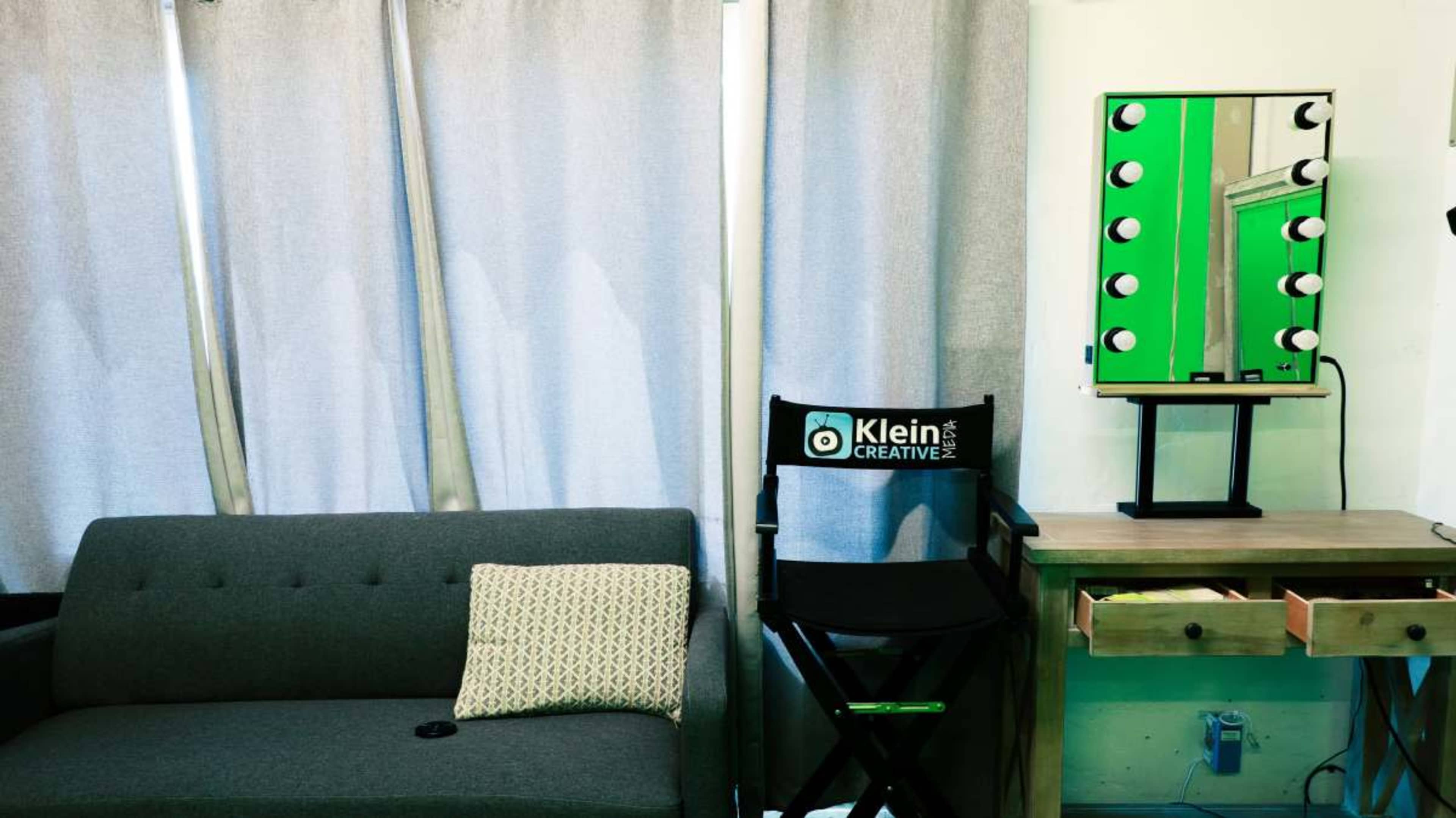 The image shows a room featuring a gray couch, a director's chair branded "Klein Creative," and a lighted mirror on a small table.