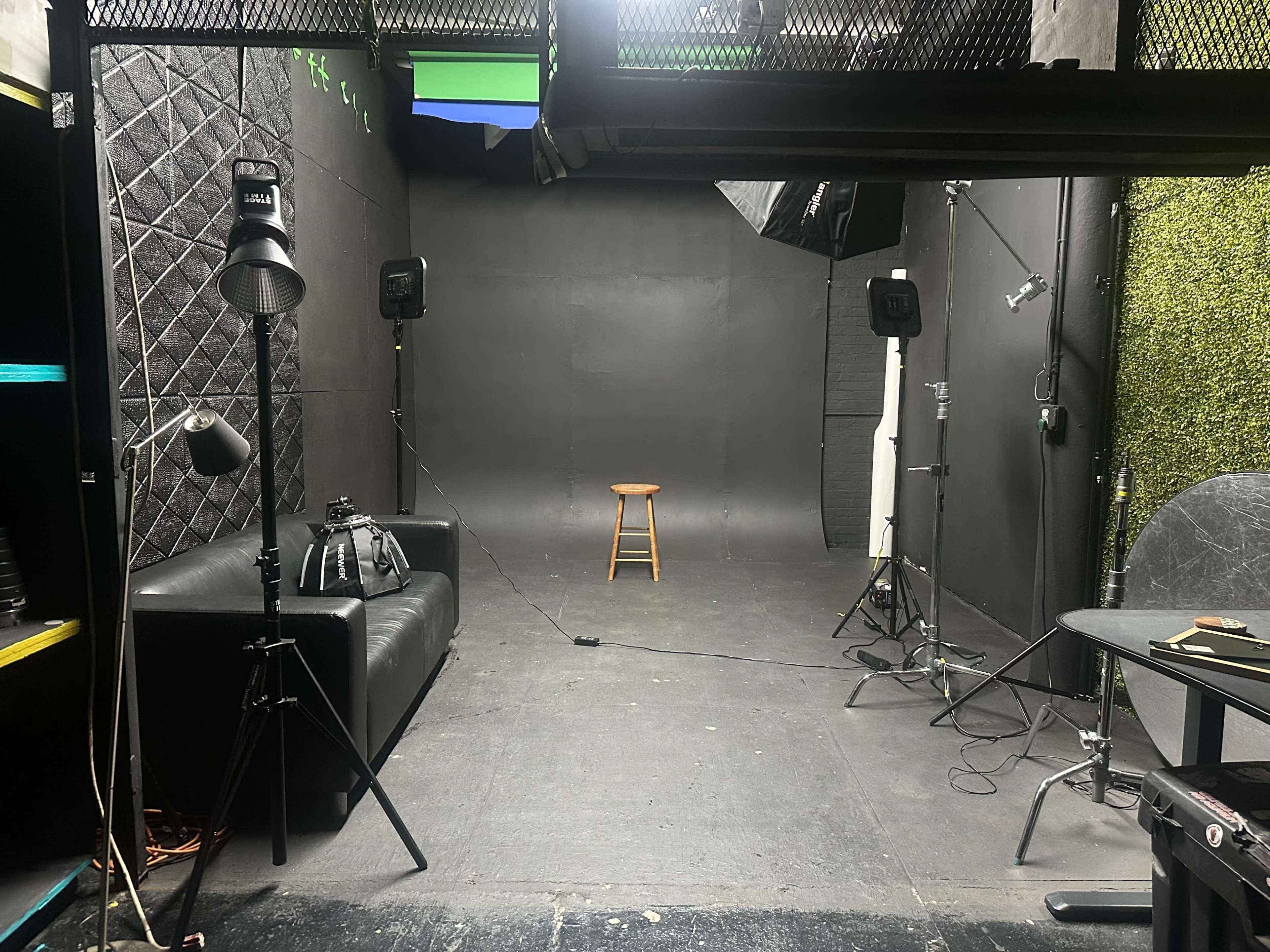 The image shows a photography studio with a black backdrop, a wooden stool in the center, and lighting equipment on either side.