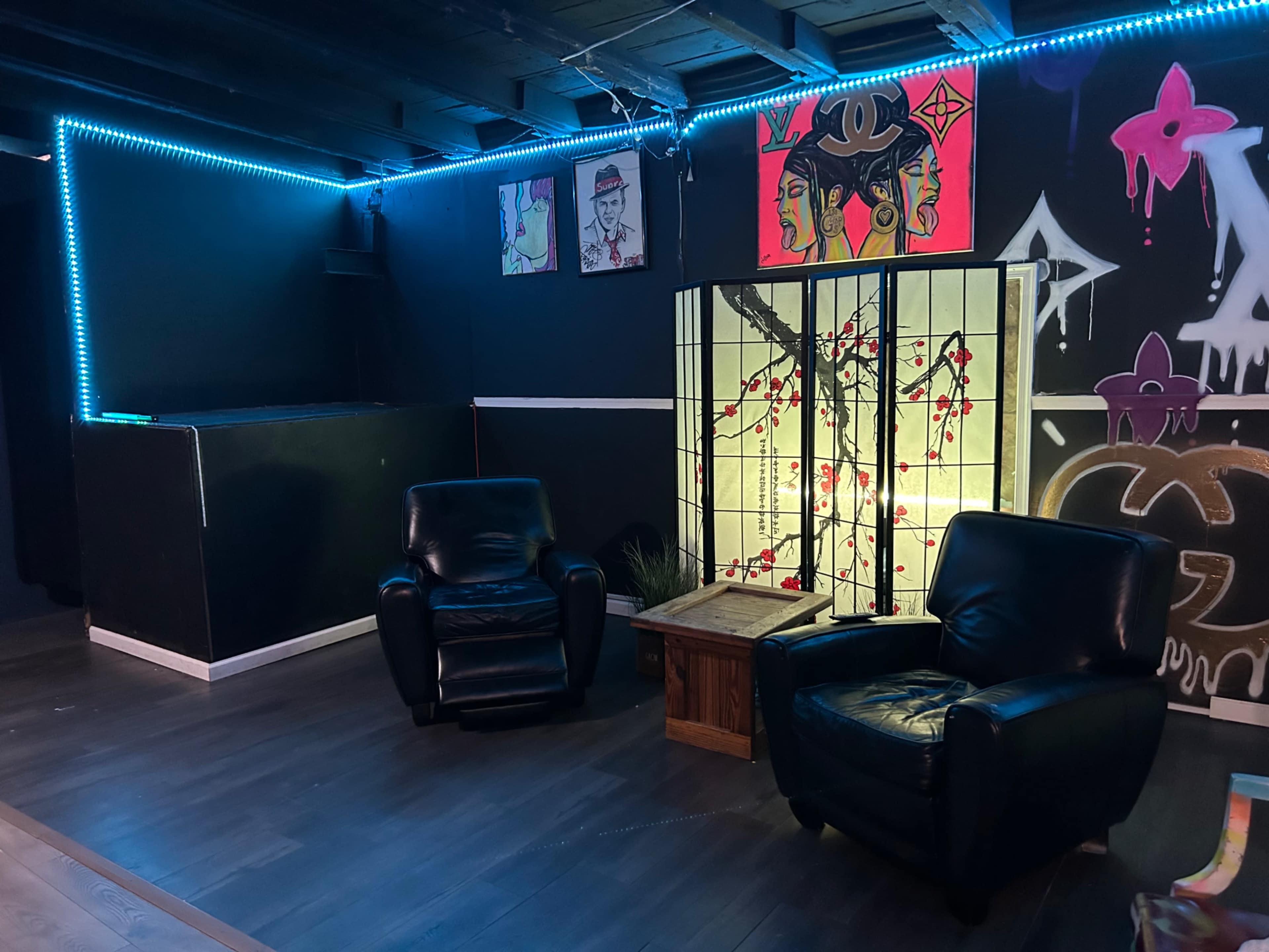 The image shows a dimly lit lounge area featuring two black leather chairs, a wooden table, and a decorative screen illuminated by neon lights, with colorful artwork on the walls.