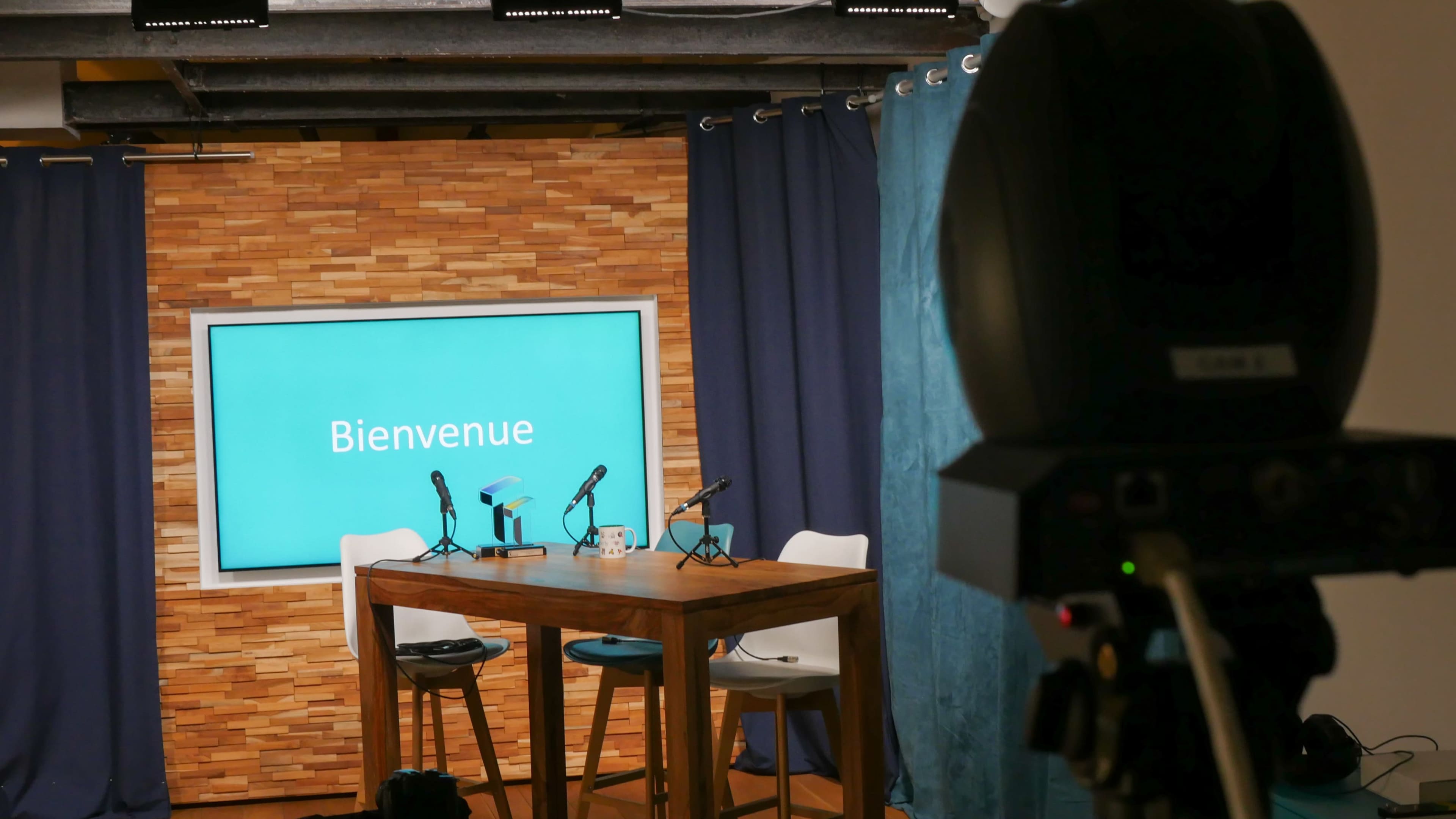 A television studio is set up with a wooden table, two microphones, and a screen displaying the word "Bienvenue," against a backdrop of wooden panels and blue curtains.
