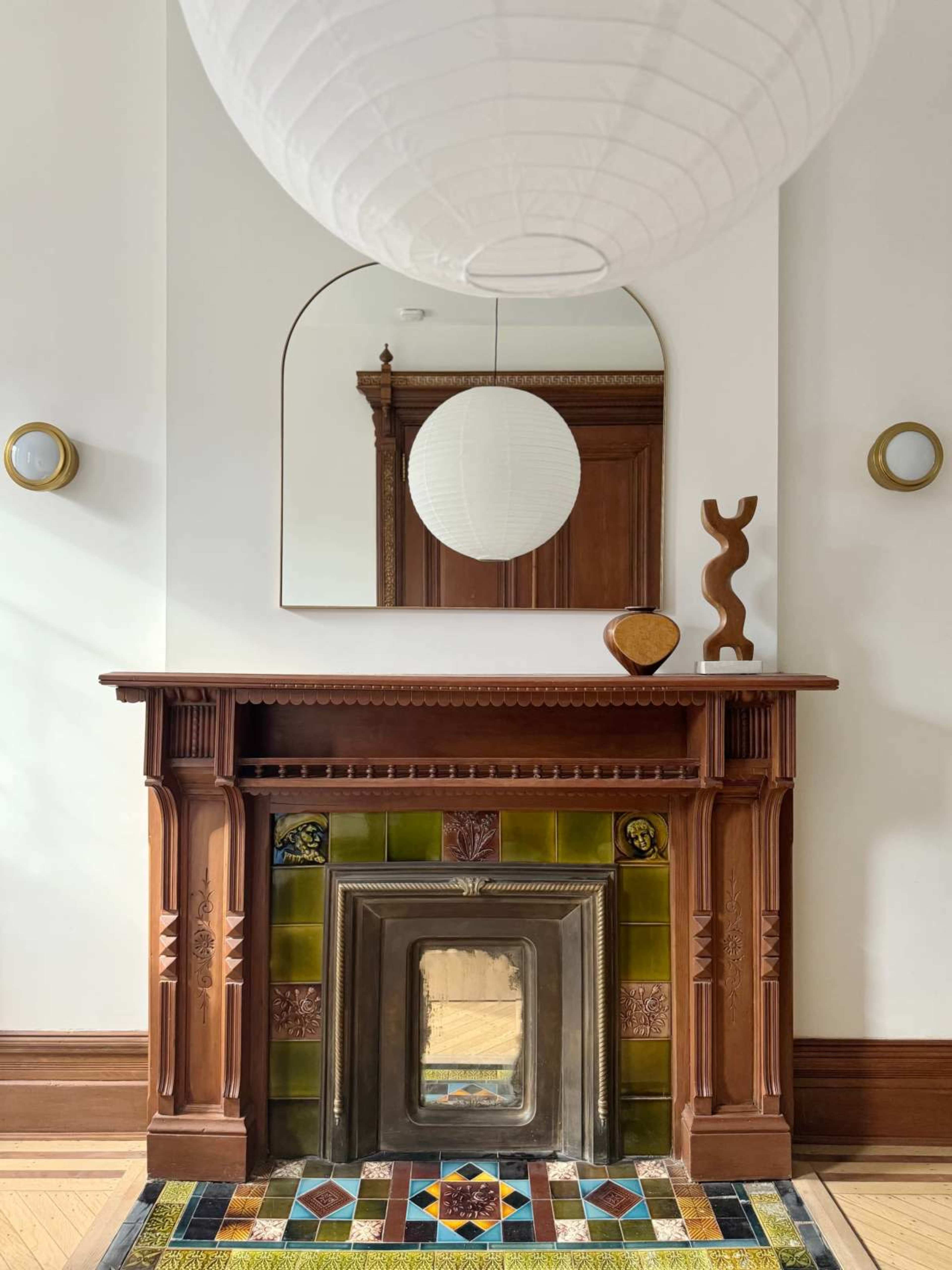 The image shows a wooden fireplace with intricate carvings, a decorative tiled hearth, and a round mirror above it, accompanied by a paper lantern light fixture and a wooden sculpture on the mantel.