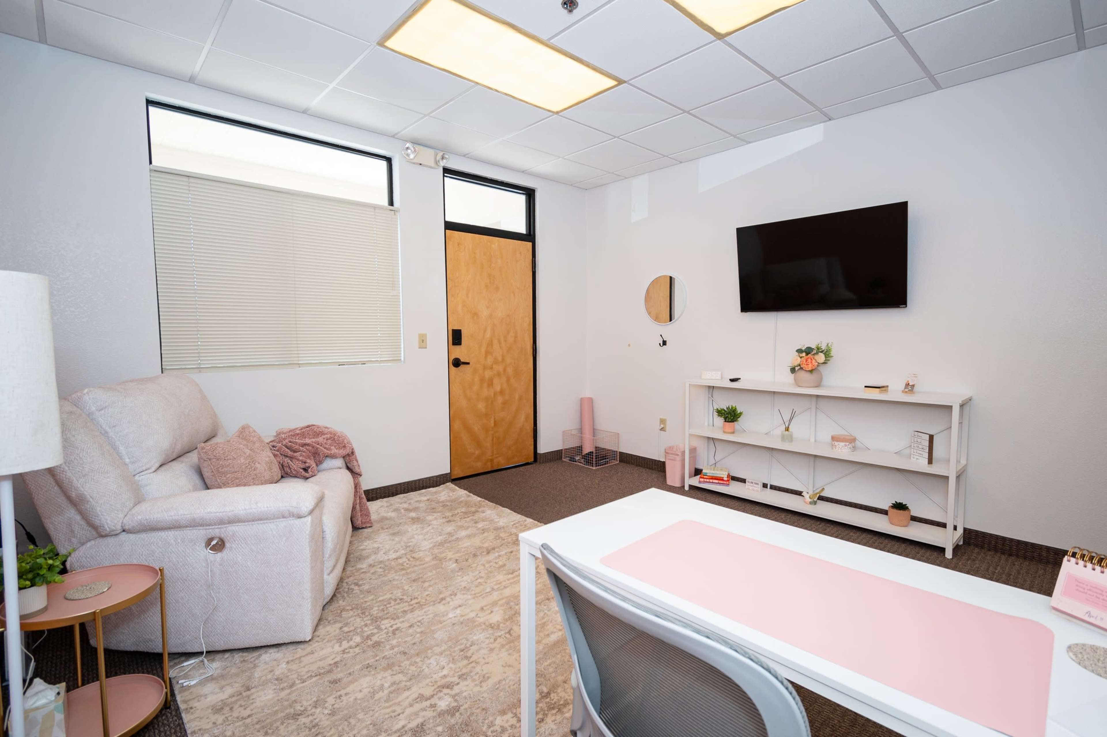 The image depicts a well-lit office space furnished with a light-colored sofa, a desk with a pink tabletop, and a wall-mounted TV.