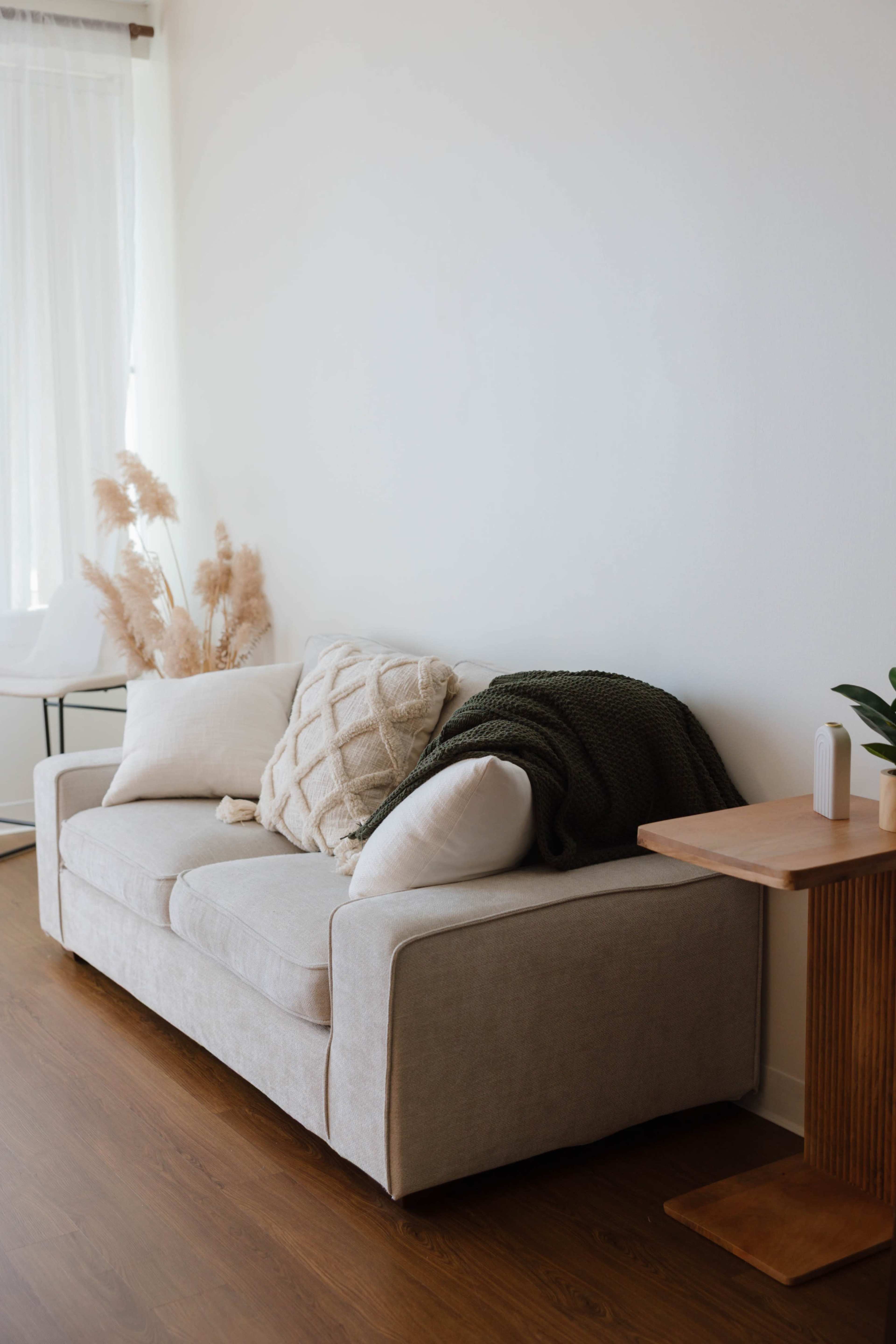 A beige couch with decorative pillows is positioned against a white wall, alongside a wooden side table and dried plants in a vase.