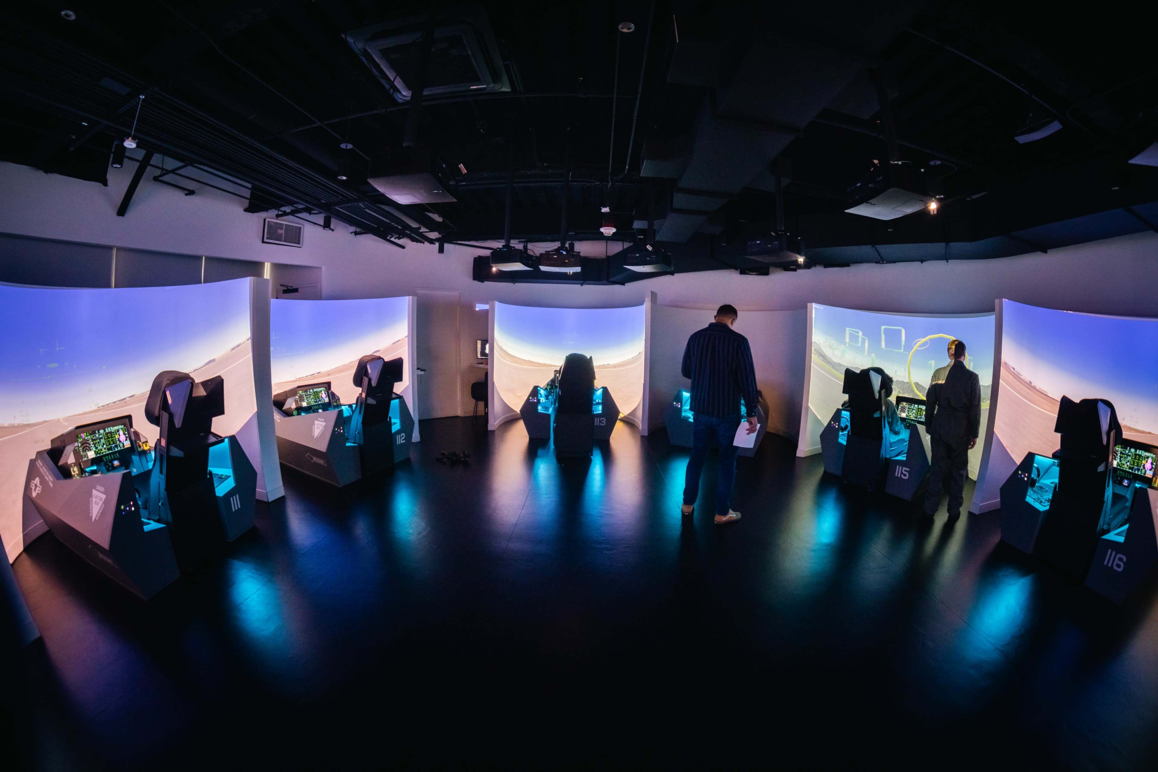 The image shows a modern simulation gaming room with multiple cockpit-style stations and large curved screens displaying a desert landscape.