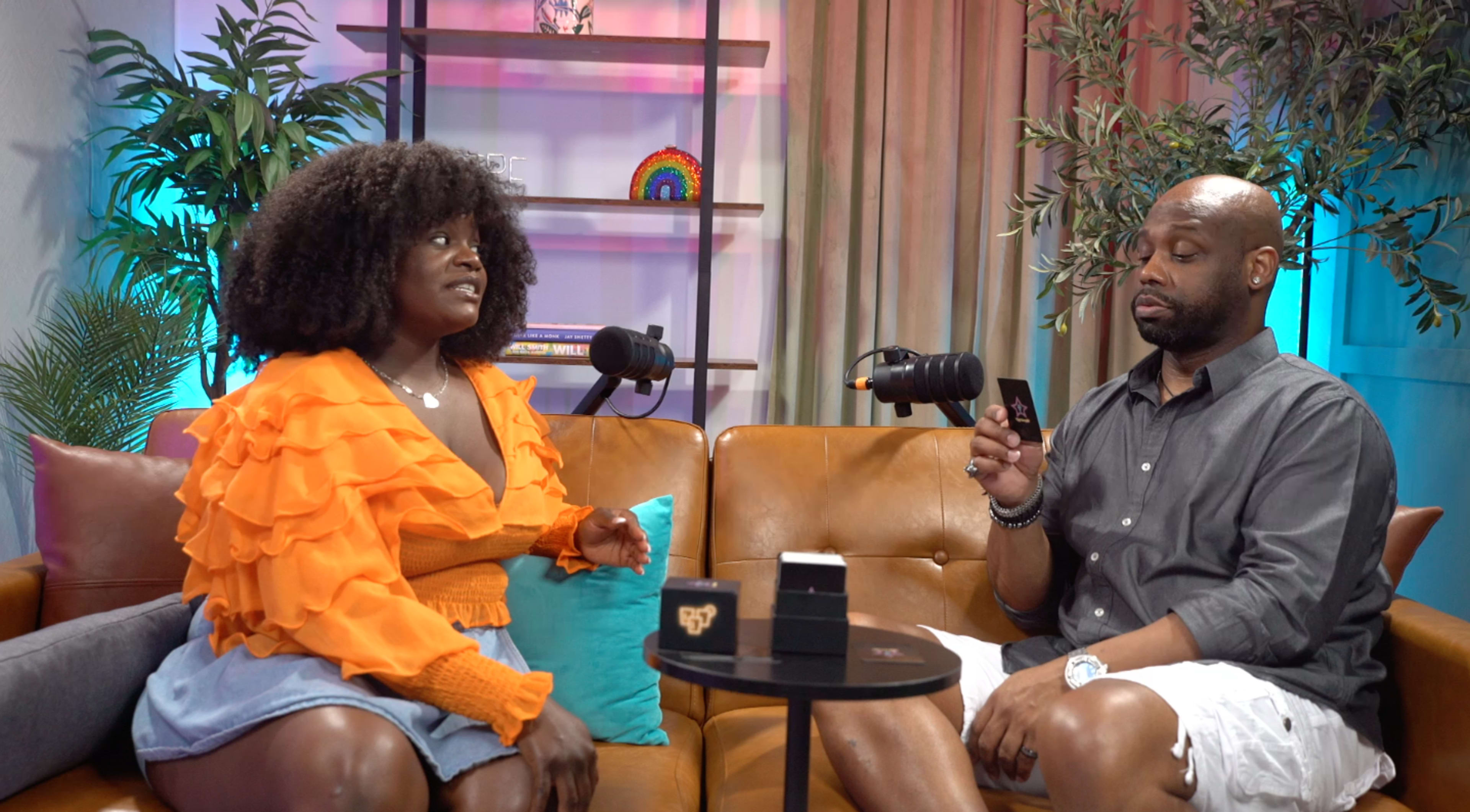 A woman in an orange sweater and a man in a gray shirt sit on a couch with microphones, discussing topics in a brightly lit studio.