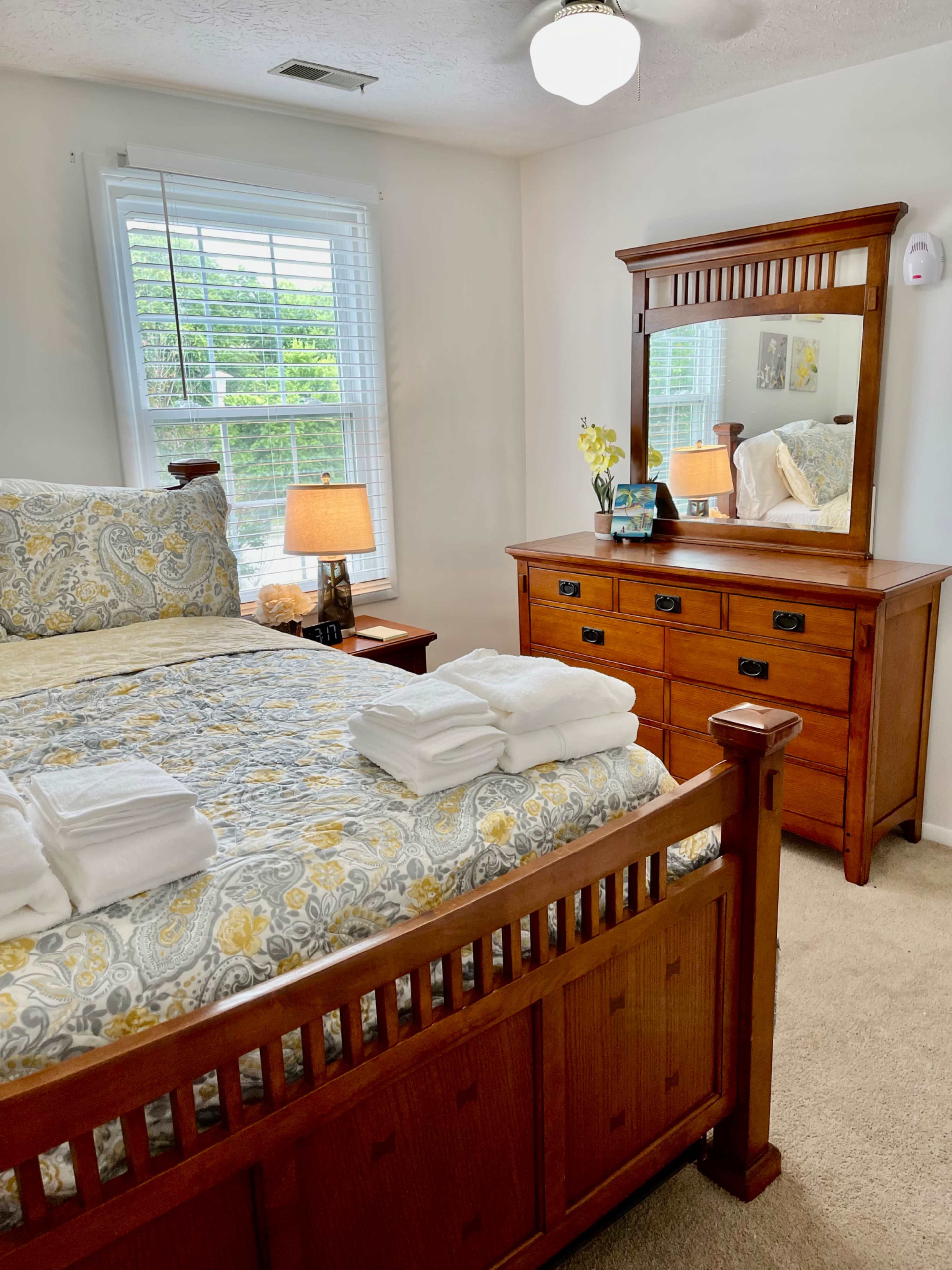 A neatly arranged bedroom features a wooden bed with a floral patterned comforter, a dresser with a mirror, and a lamp on a bedside table.