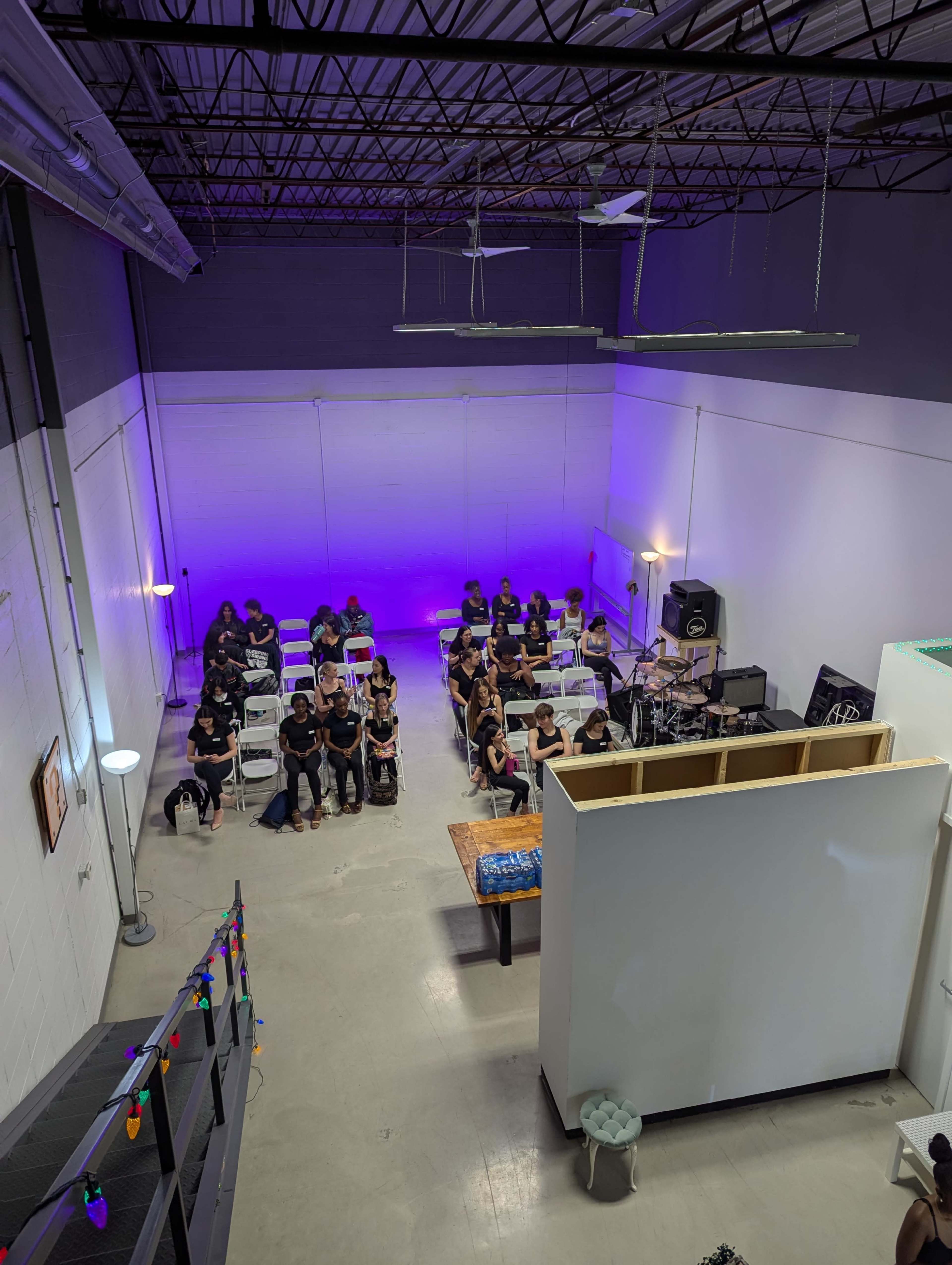A group of people in black shirts sits at tables in a well-lit room with purple lighting and a staircase in the foreground.