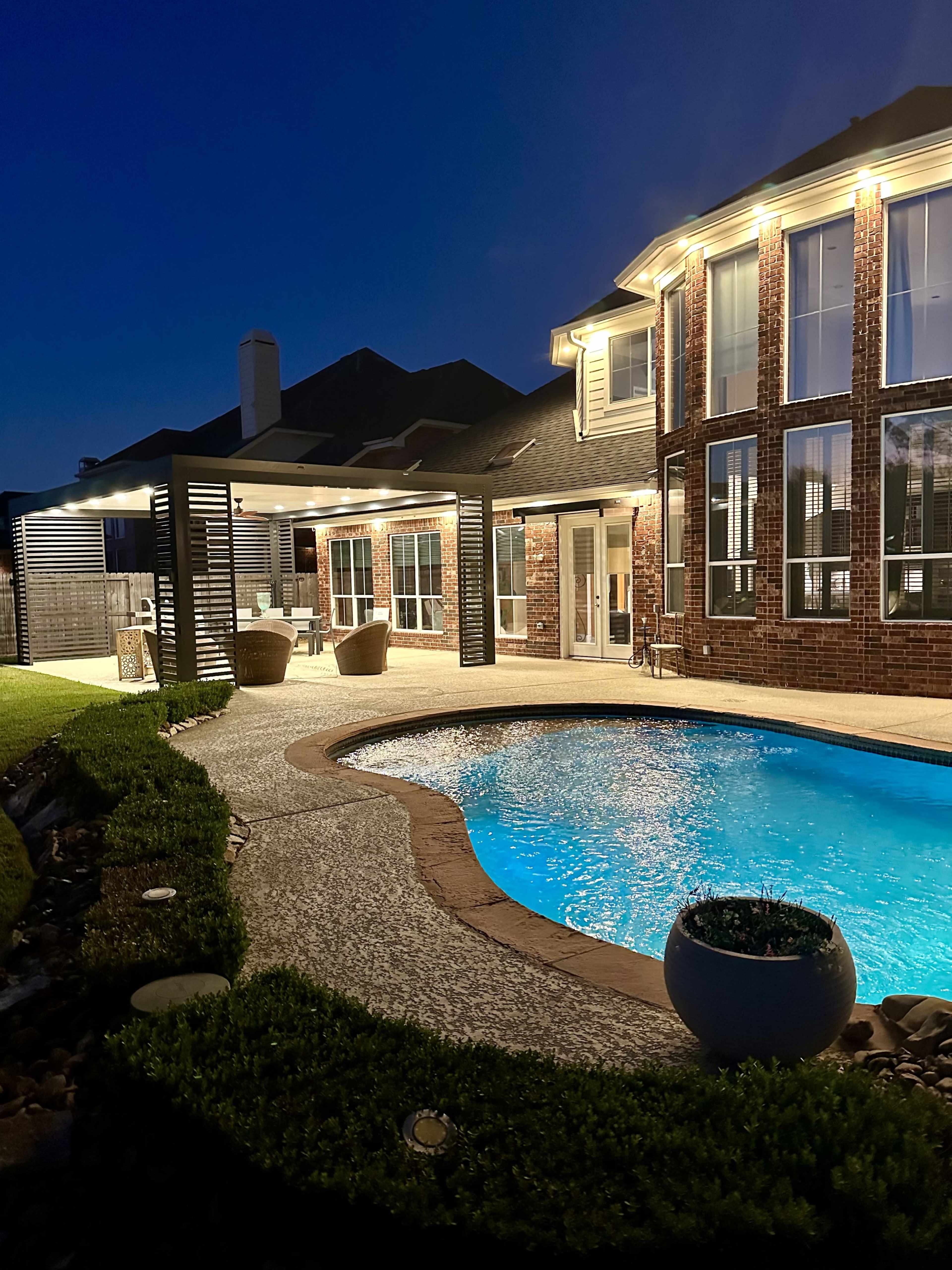 A well-lit backyard features a swimming pool surrounded by a patio and a brick house with large windows.