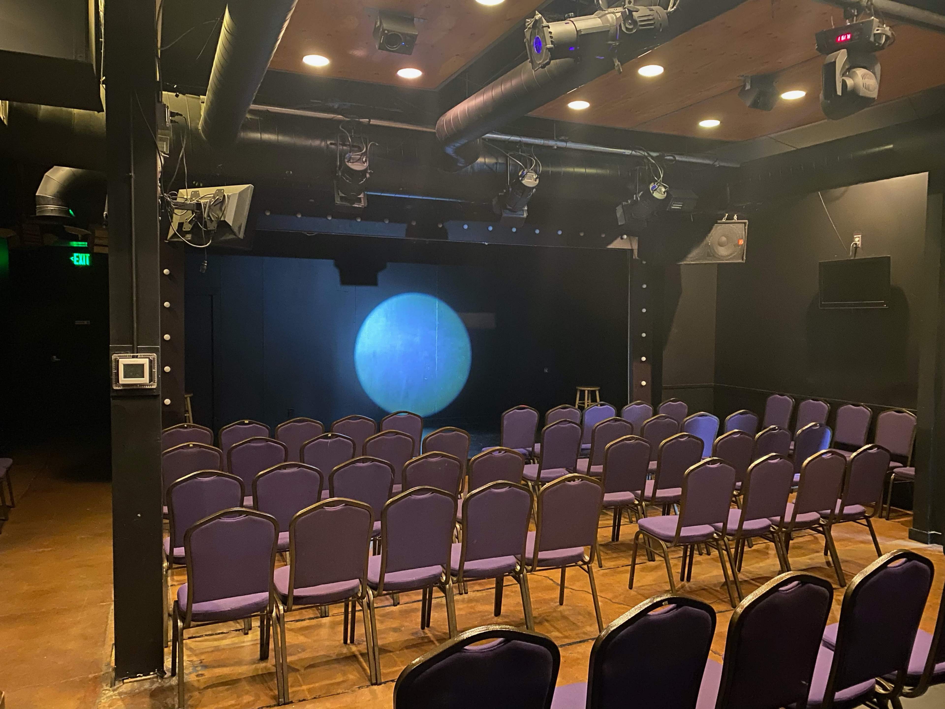 The image shows a small theater space with rows of purple chairs facing a dimly lit stage featuring a circular light projection.