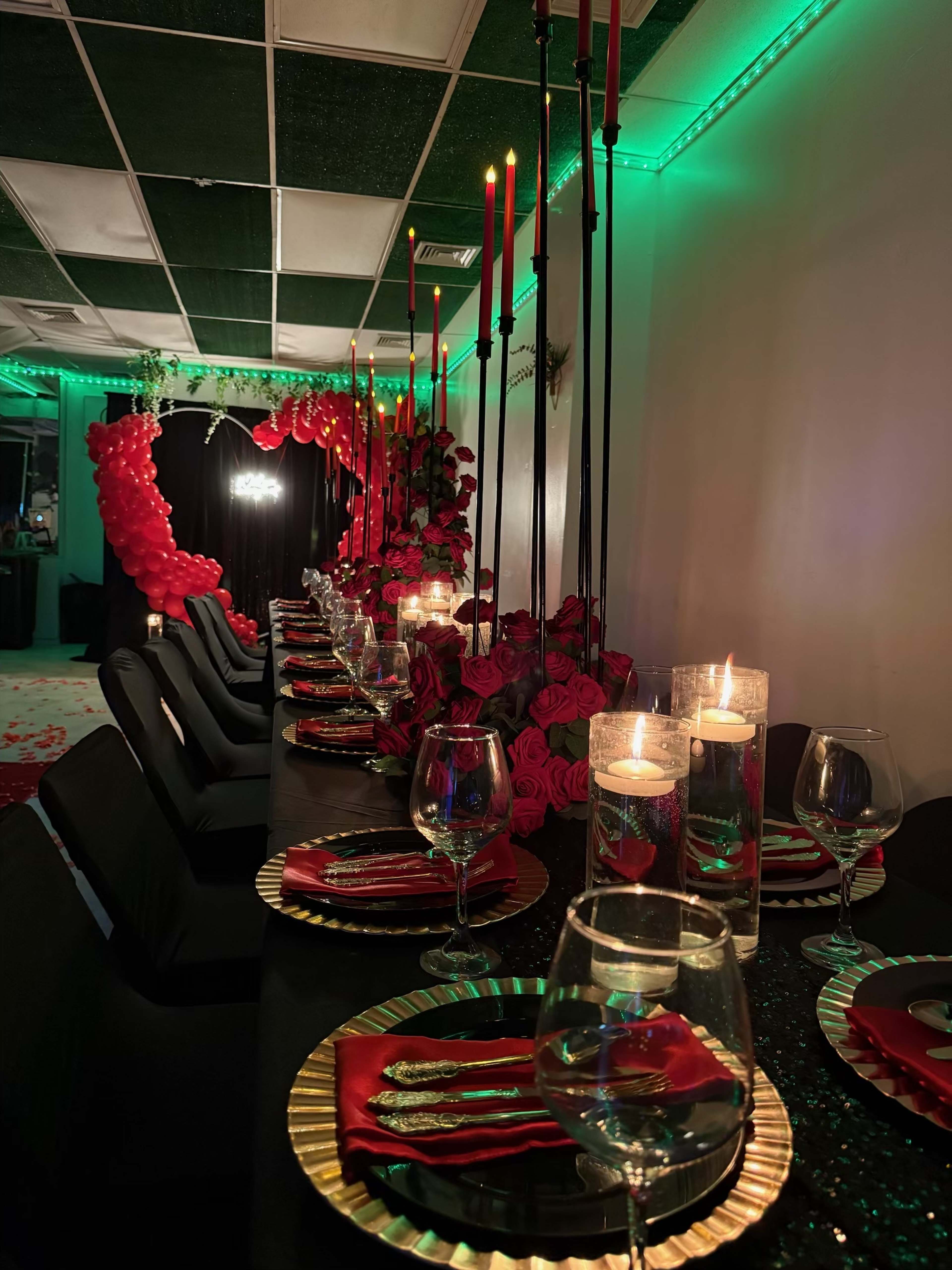 A long dining table is elegantly set with red napkins, candles, and black chairs, surrounded by decorations of red balloons and flowers, illuminated by green lighting.