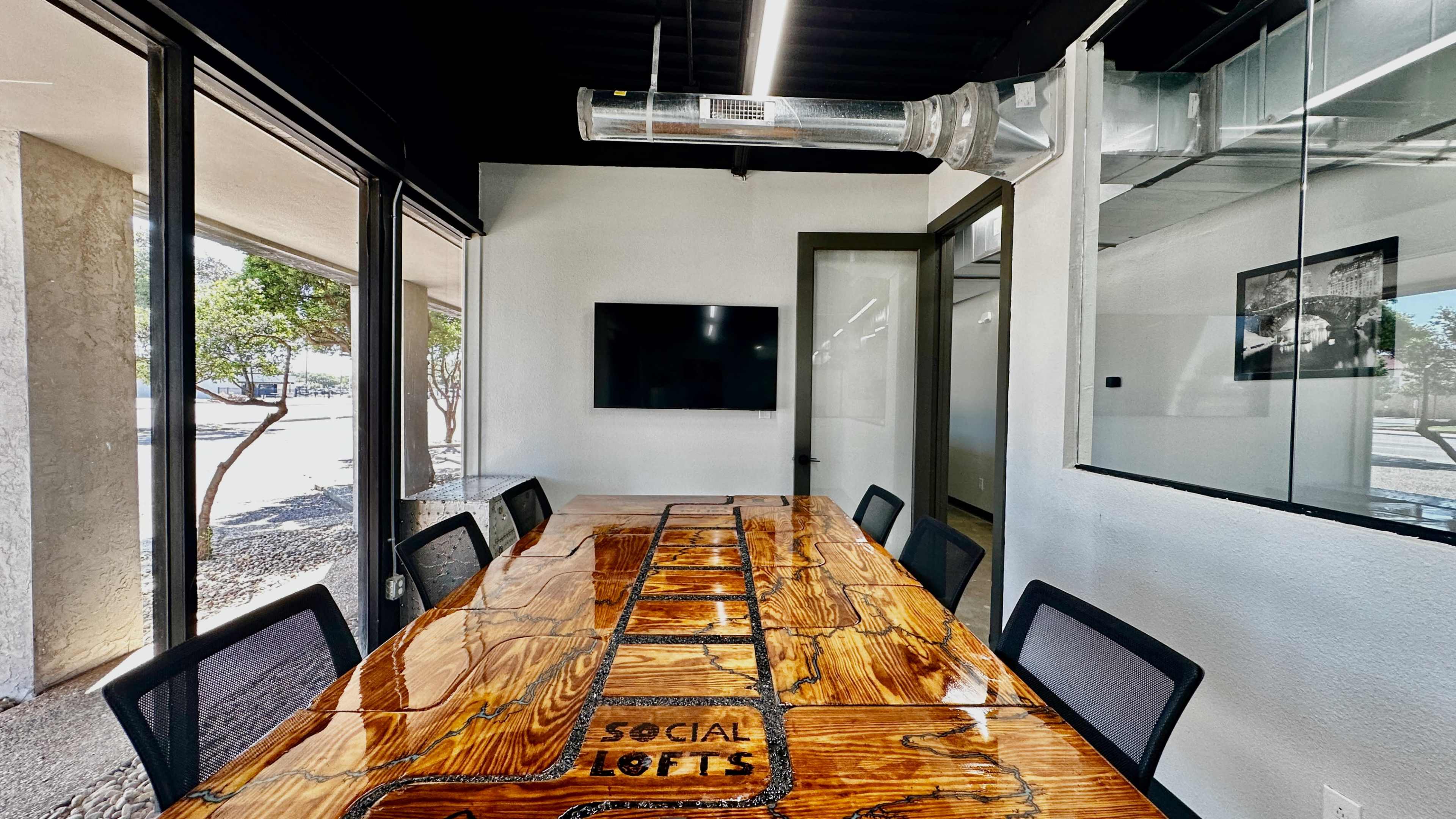 A modern conference room features a large wooden table with "SOCIAL LOFTS" engraved in its center, surrounded by black chairs and glass walls.