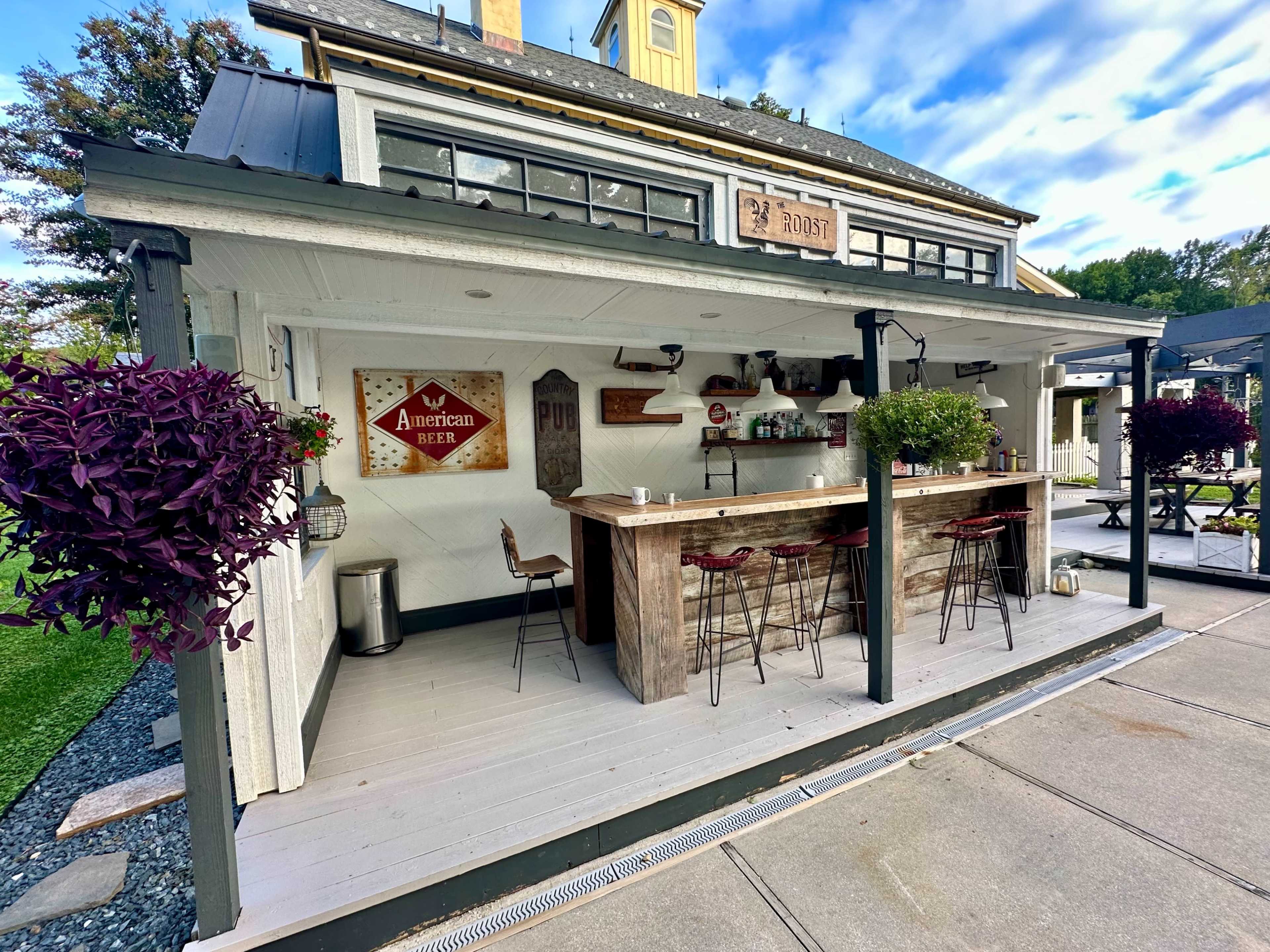 The image shows a spacious outdoor bar area with wooden furniture, planters, and a sign featuring "American Beer" attached to the wall.