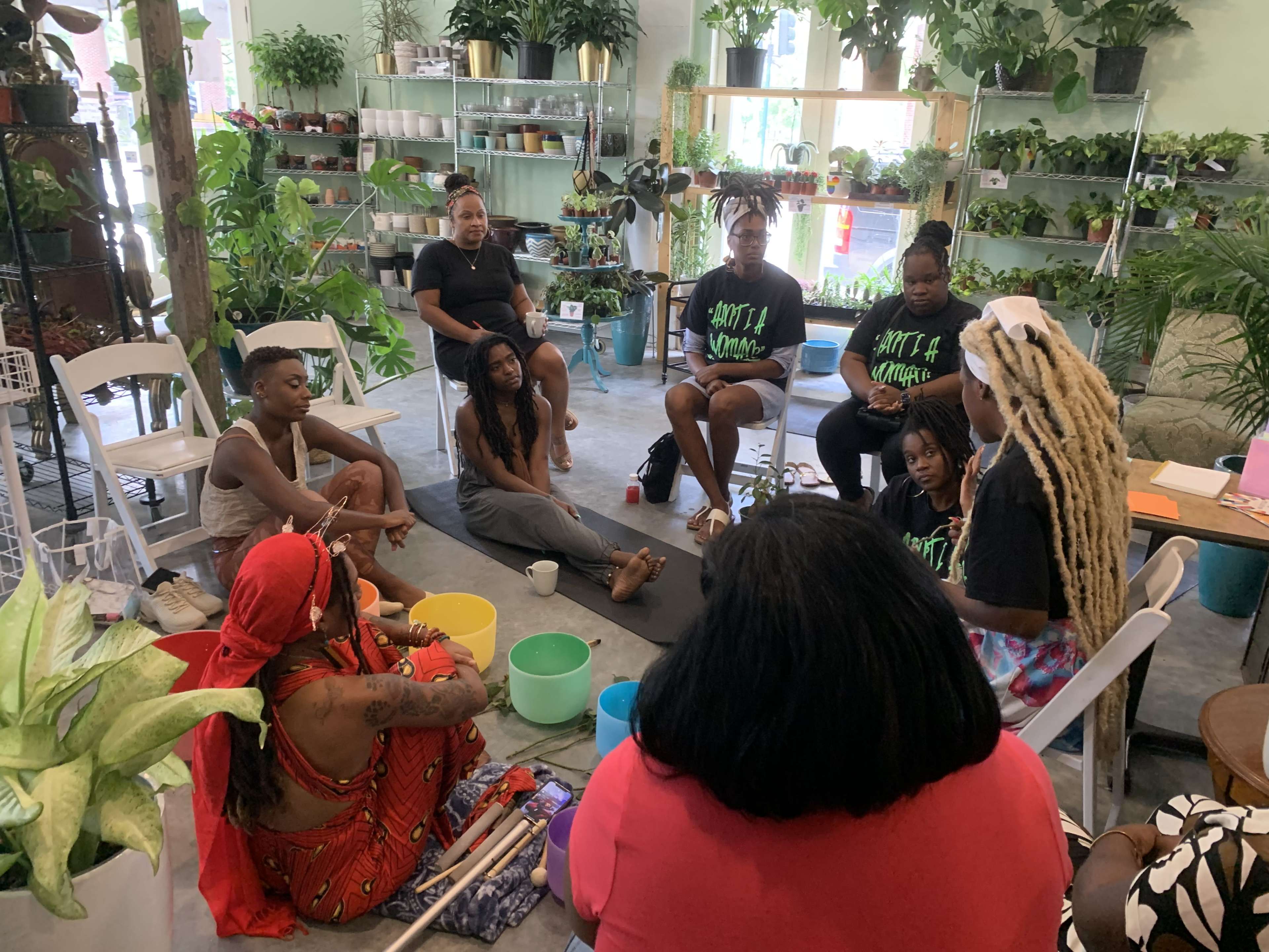 A group of people sits in a circular arrangement in a plant-filled room, engaged in a discussion or activity.