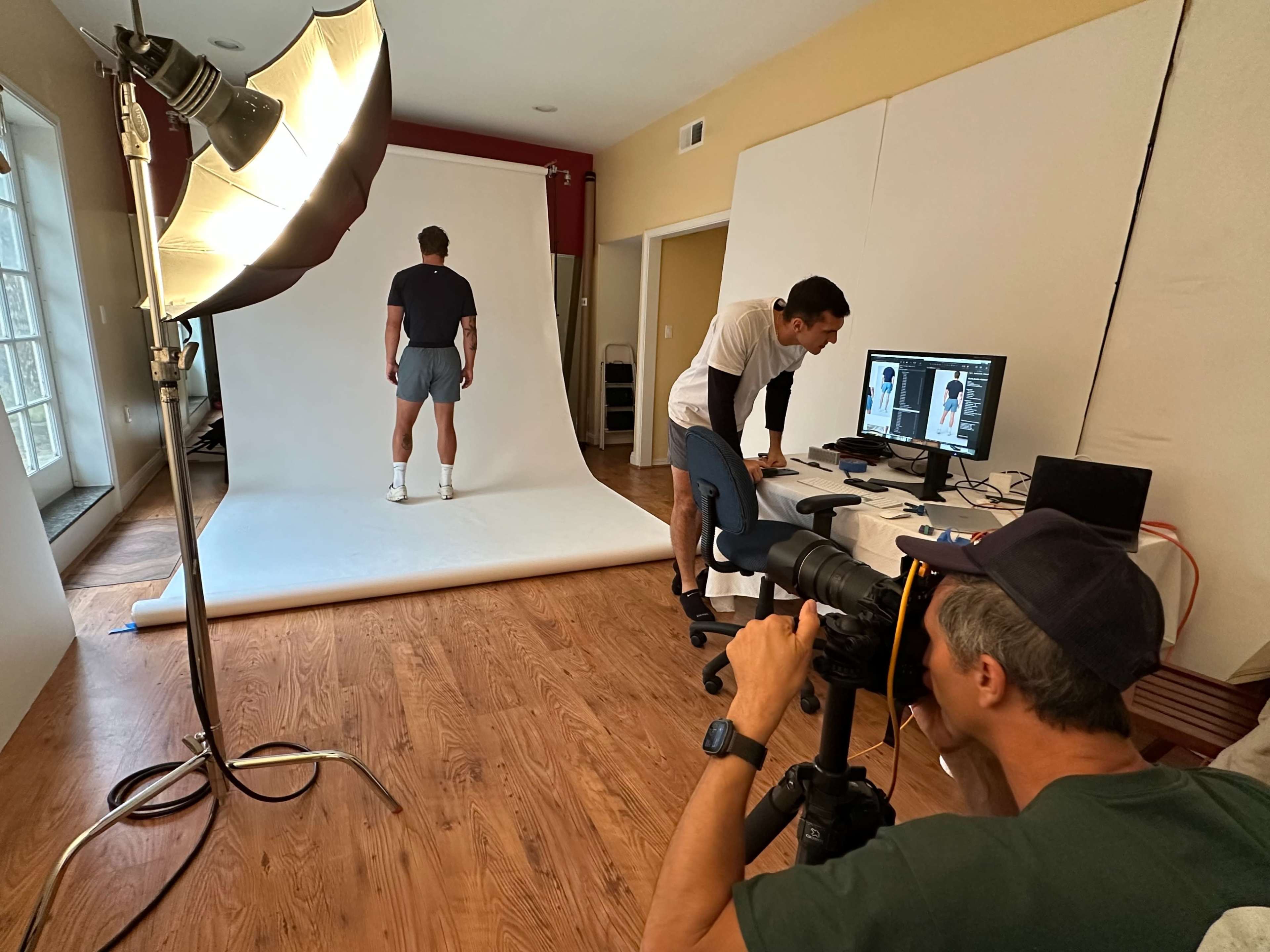A photographer captures a model posing against a white backdrop in a studio, while a second person reviews images on a computer nearby.