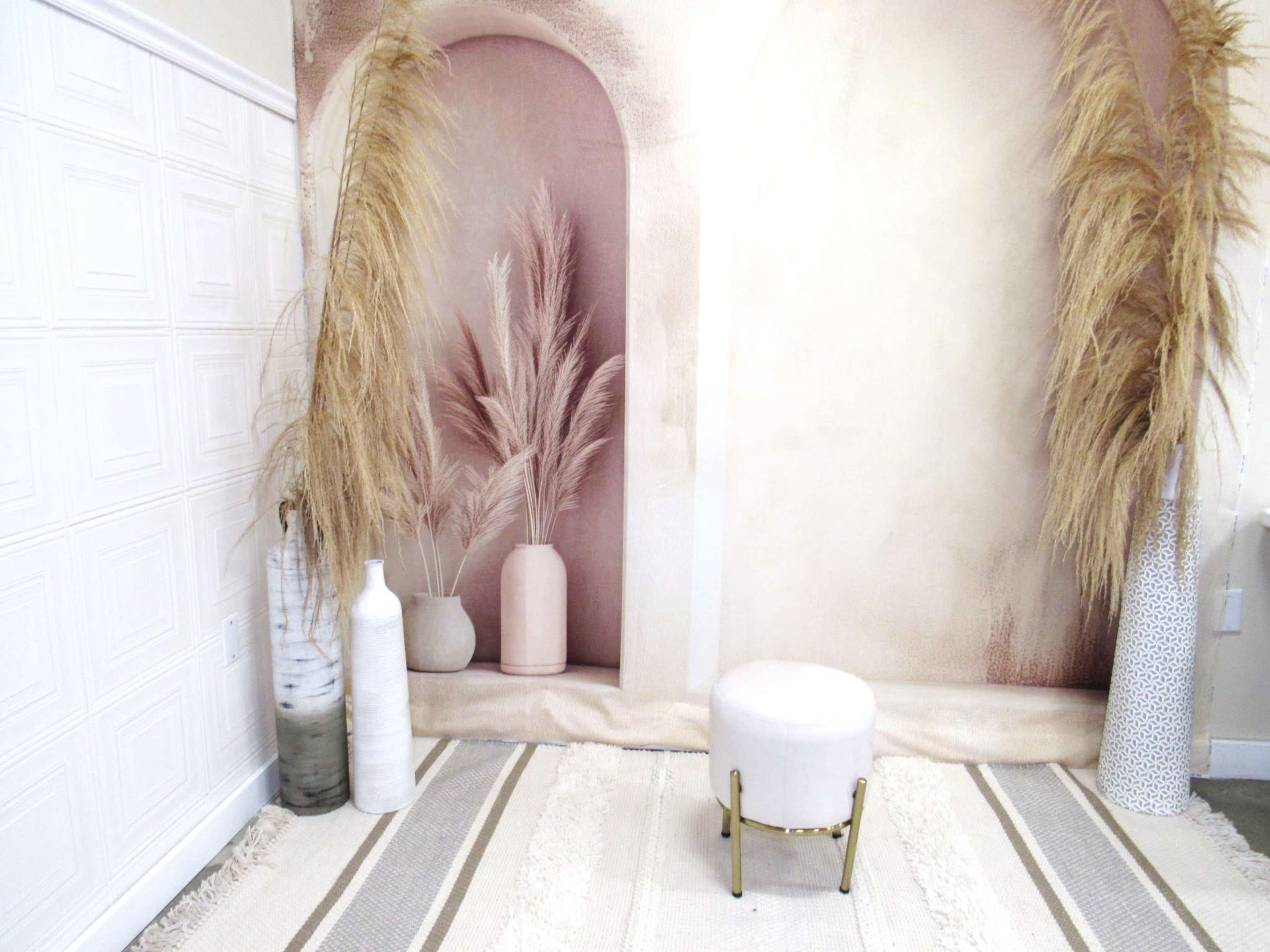 A styled interior scene featuring tall dried plants in vases, a small round stool, and a textured rug against a backdrop with arched shapes.